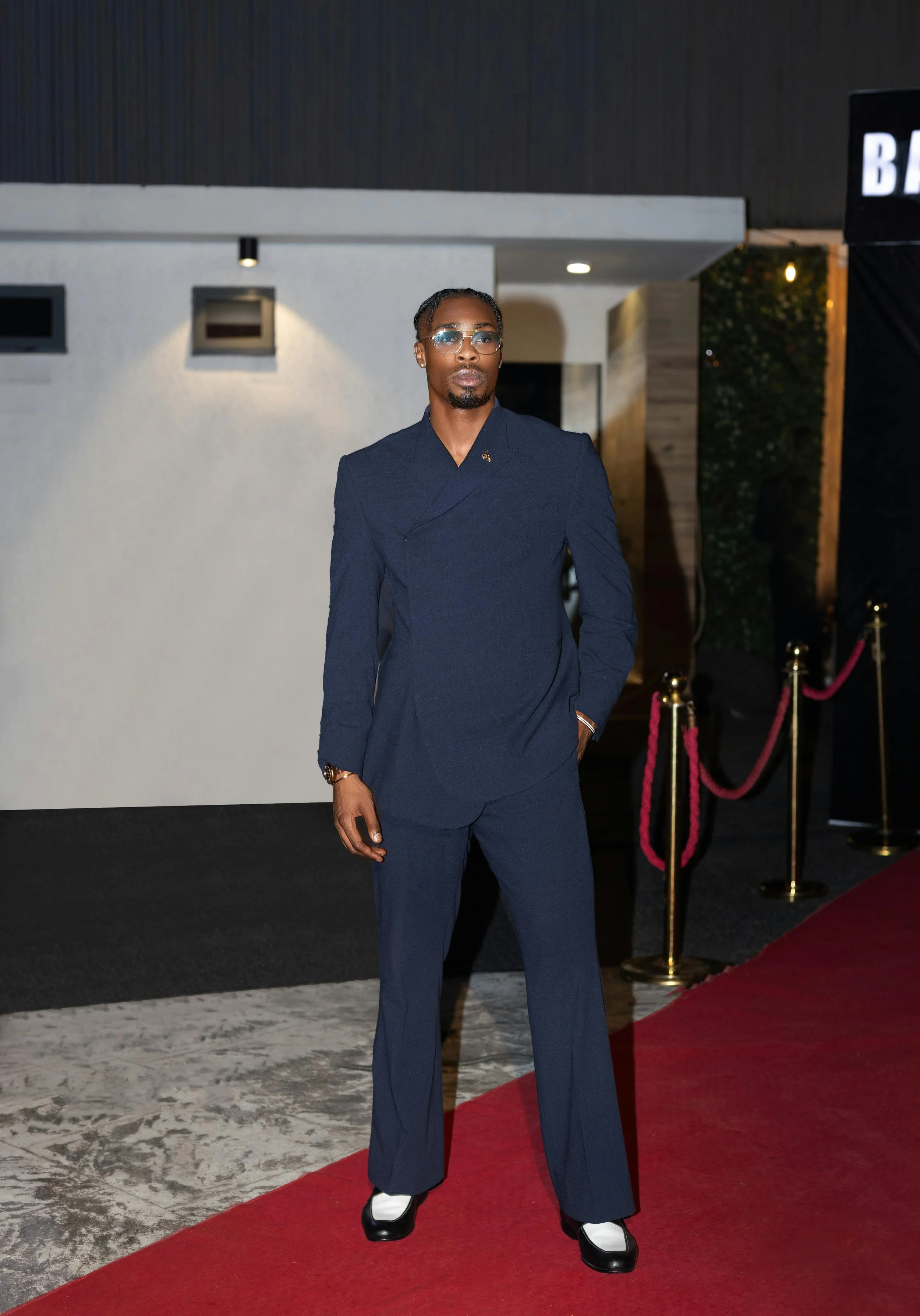 A man dressed in a navy blue suit with a double-breasted jacket and wide-leg trousers, standing on a red carpet at a formal event during the evening, with a modern building in the background.