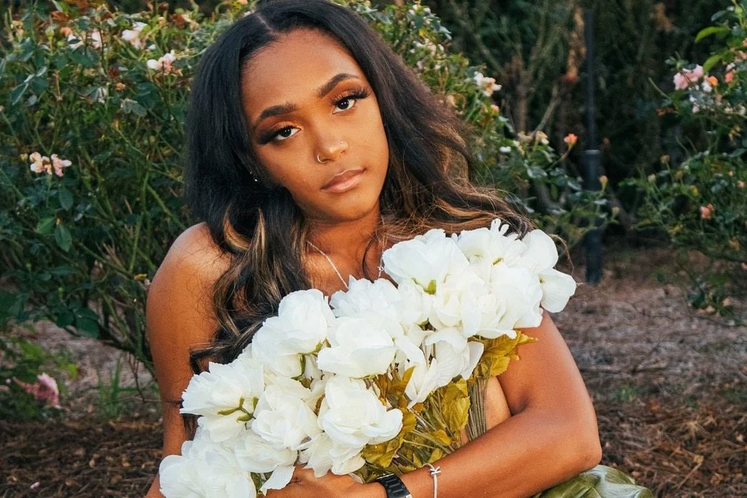 A woman with long dark hair and medium skin tone holding a bouquet of white flowers outdoors, with green foliage and pink flowers in the background.