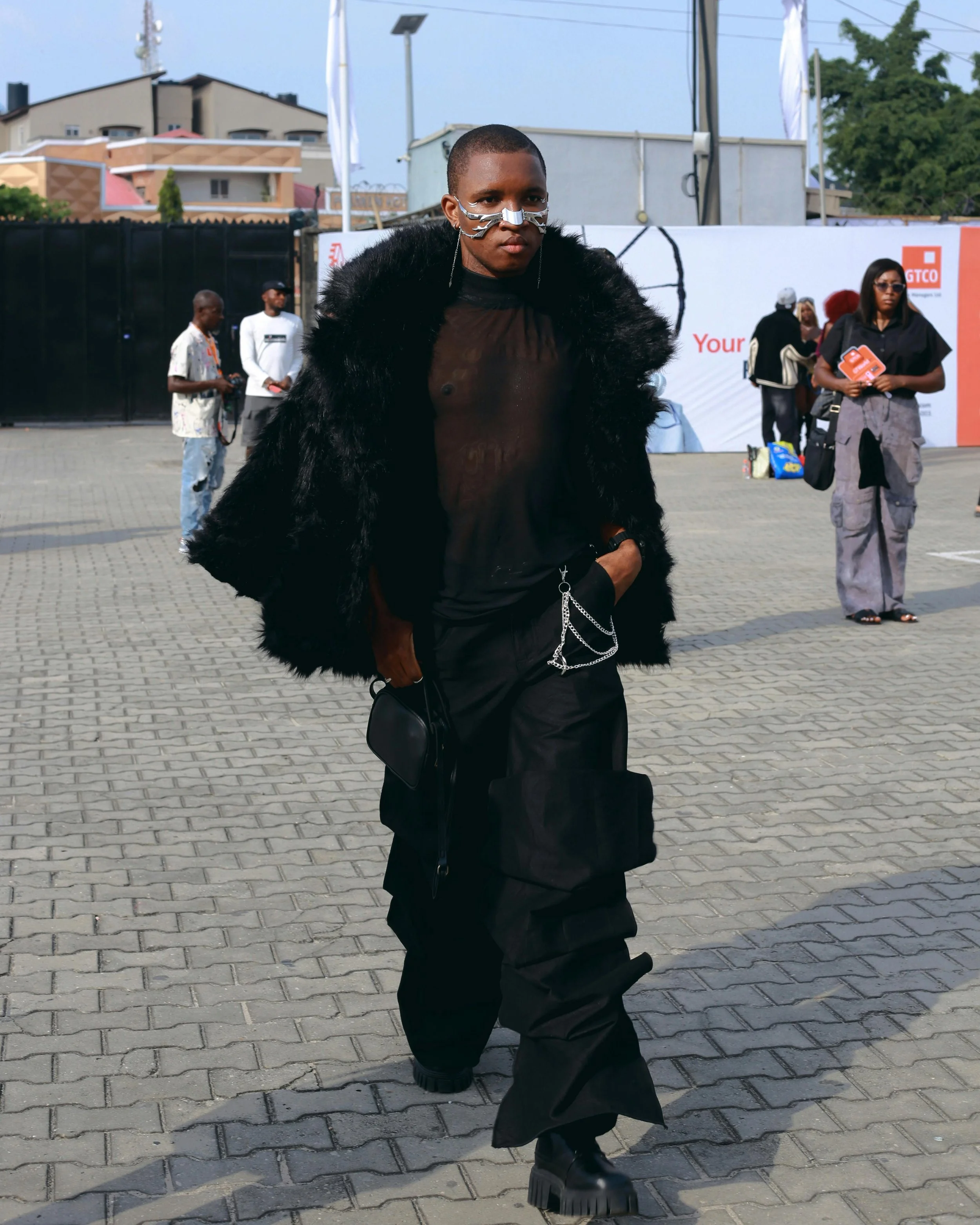 A person walking outdoors wearing a black sheer top, a black fur jacket, black cargo pants with chains, chunky black boots, and a nose chain accessory.