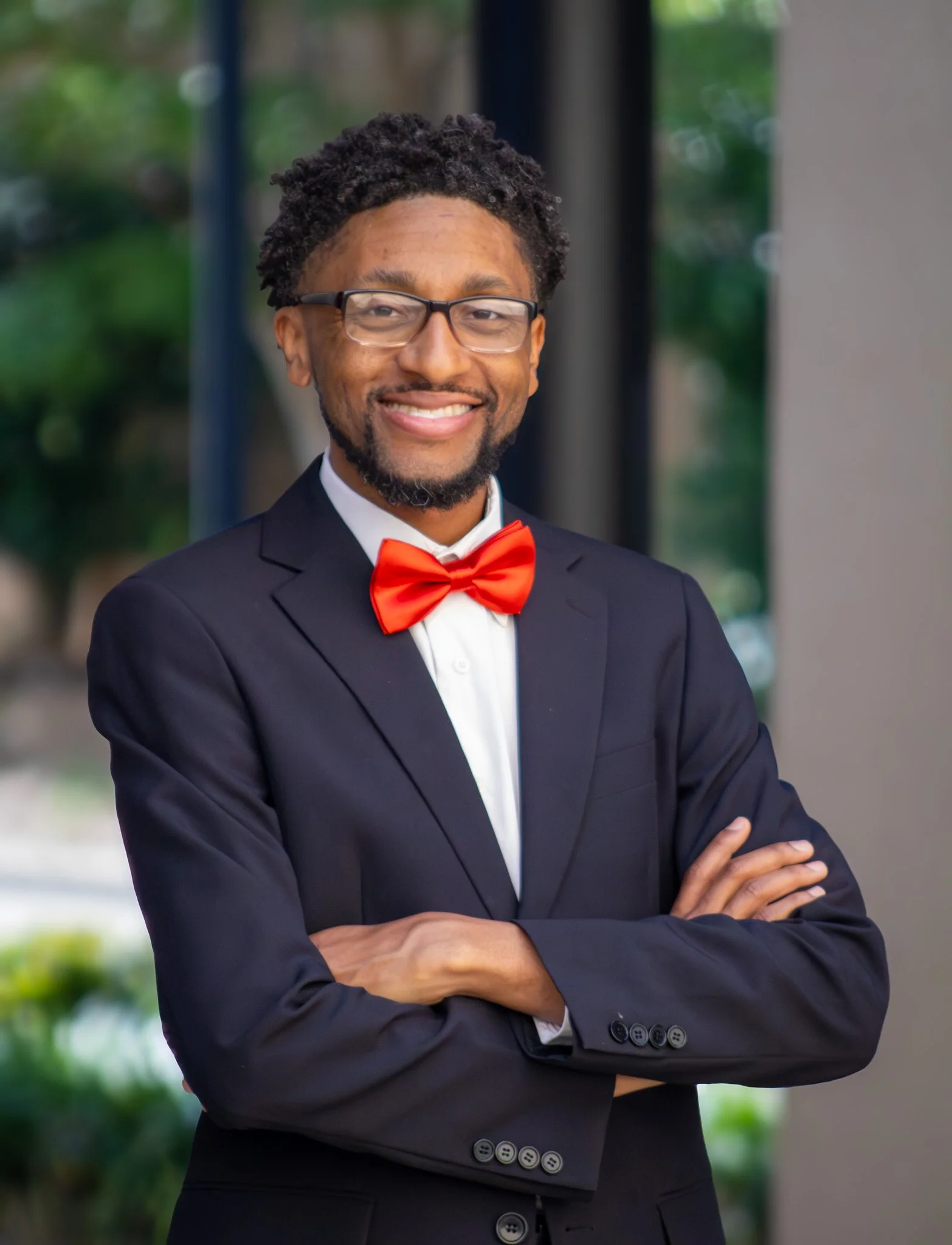 A man wearing glasses, a black suit, and a red bow tie, standing outdoors with arms crossed, smiling.