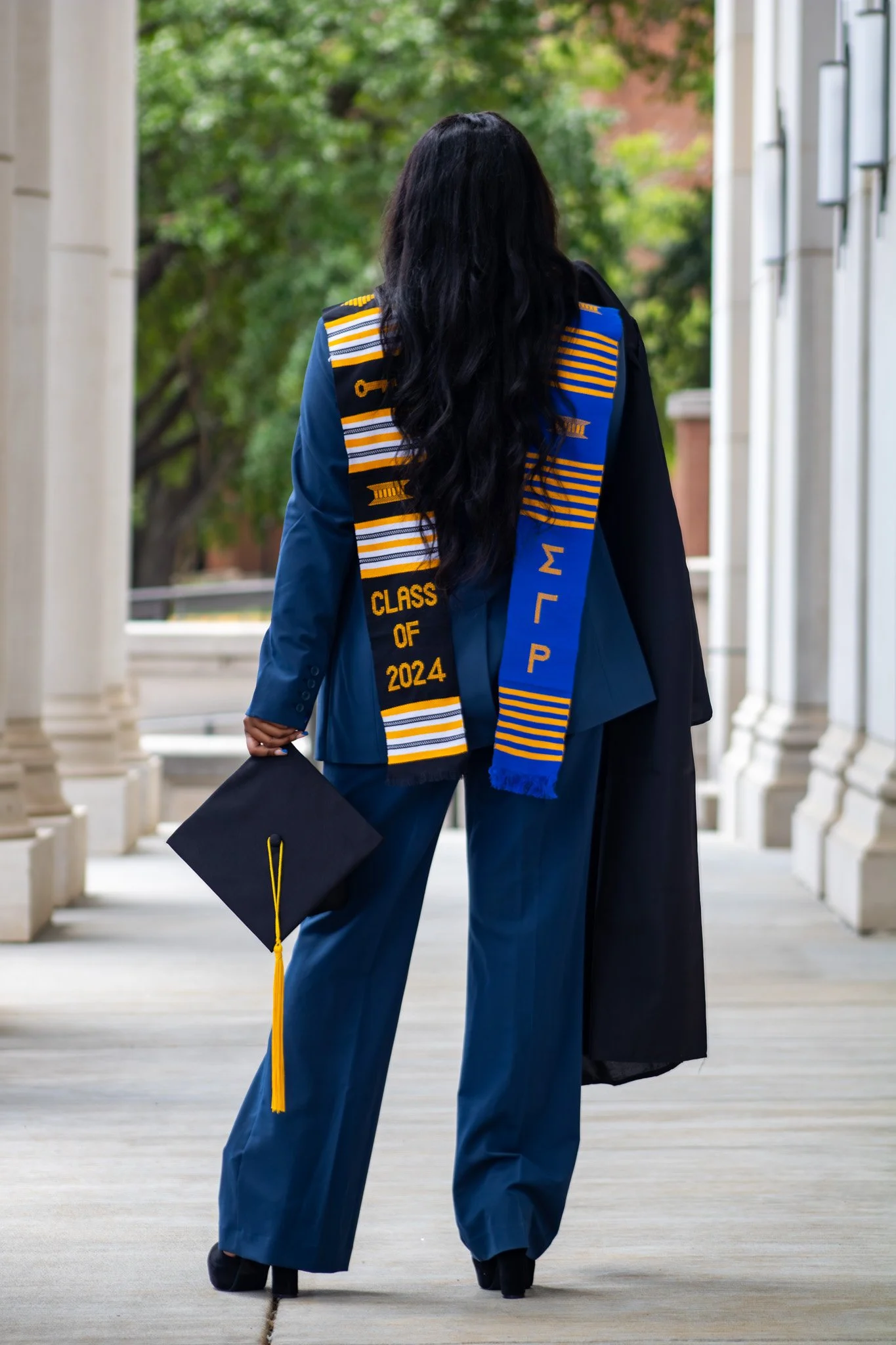 Graduation ceremony with a woman in a blue cap and gown holding a diploma, wearing a stole with 'Class of 2024' and Greek letters, standing outdoors amid trees.