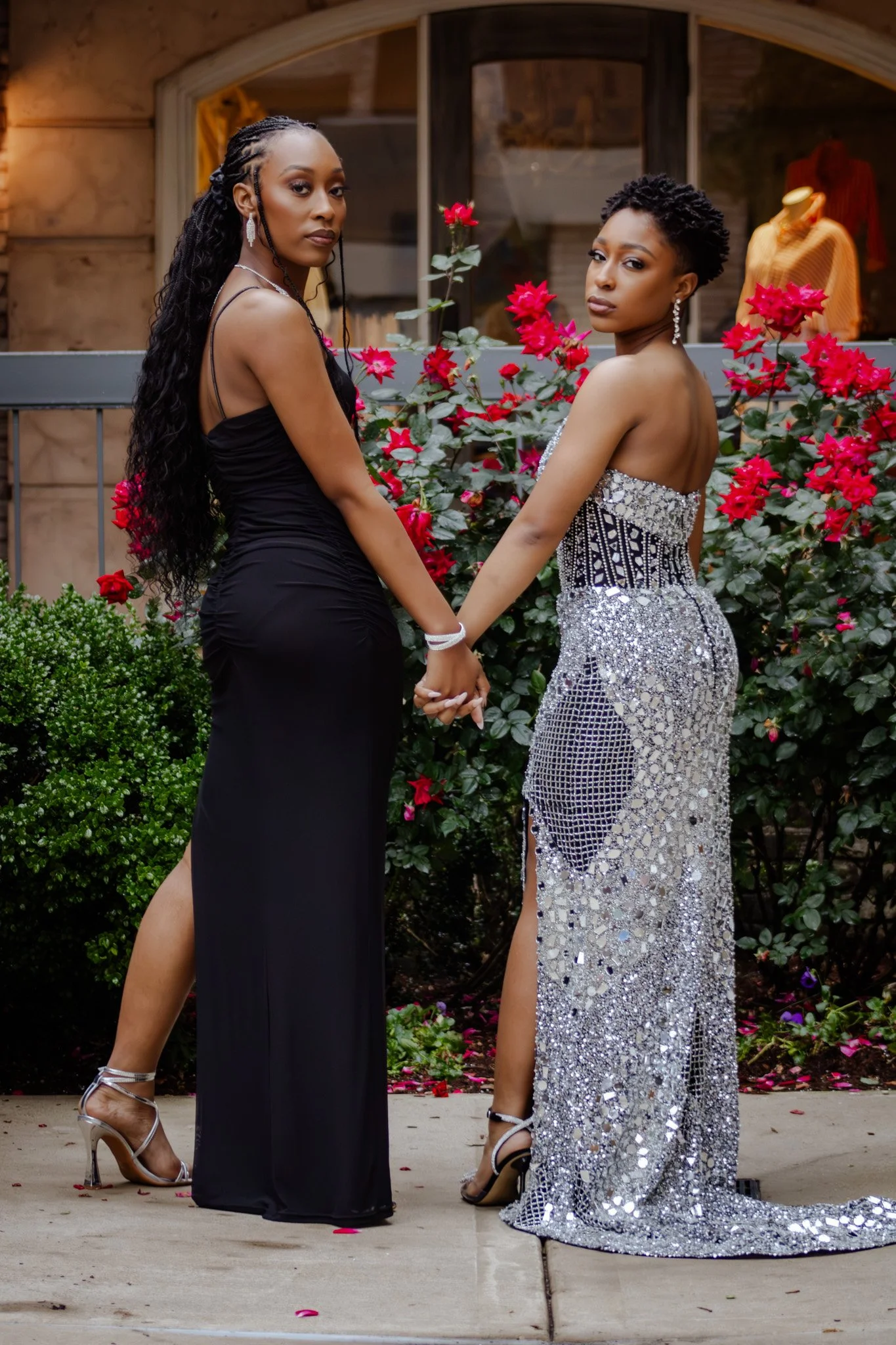Two women in evening gowns holding hands, standing outdoors in front of pink and red flowering bushes.