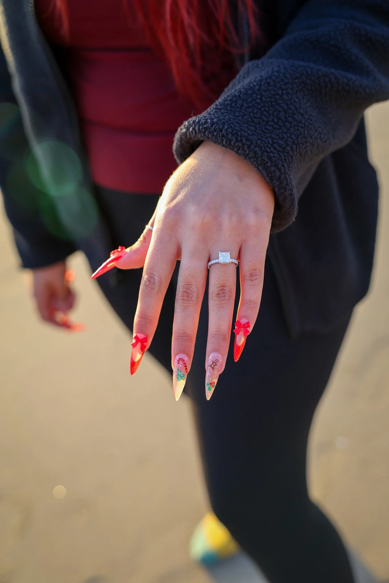 Close-up of a woman's hand showing a large diamond engagement ring and Christmas-themed nail art, with a blurred background.