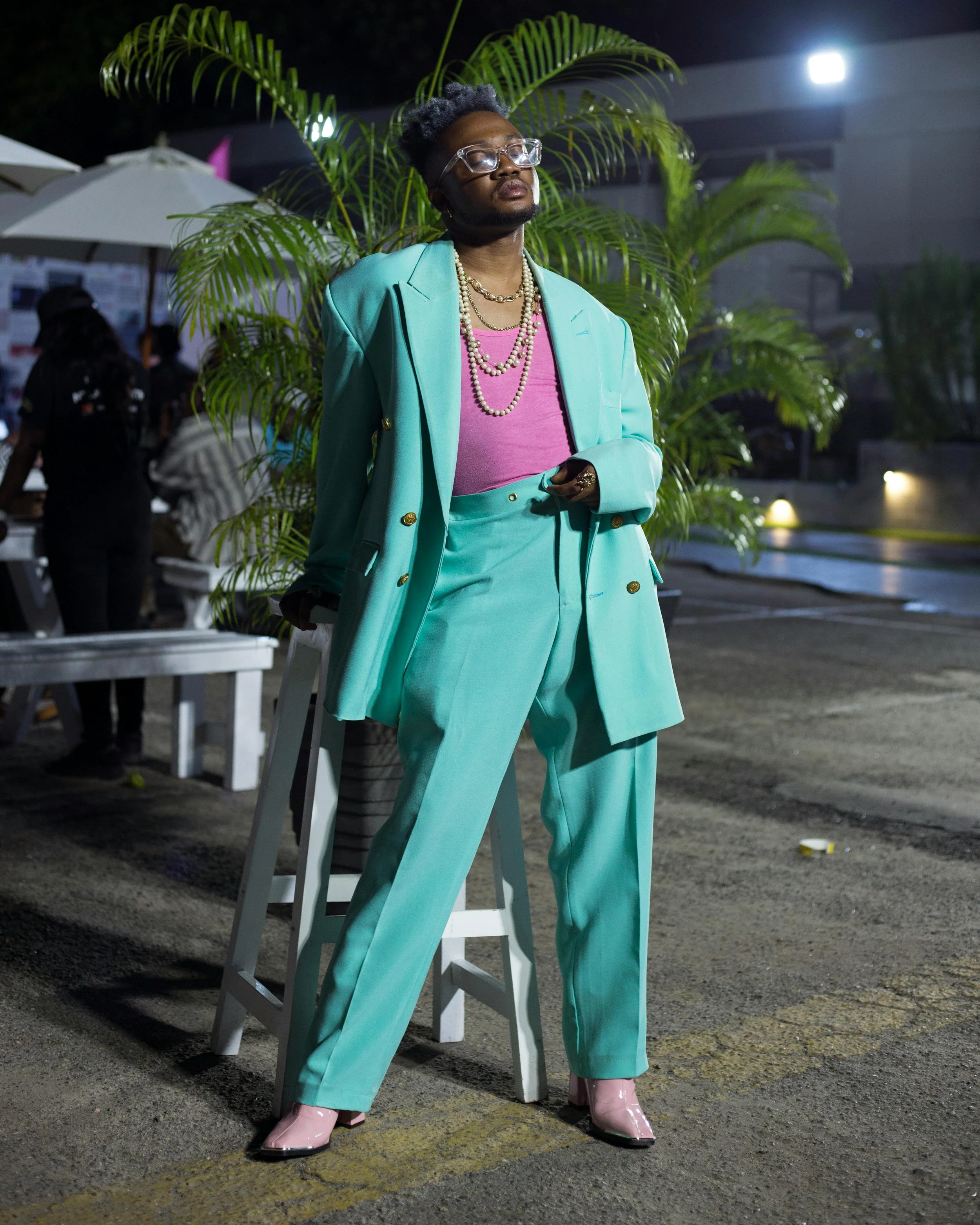 A person with short gray and black hair, wearing glasses, a turquoise suit, pink shirt, and pink shoes, standing outdoors at night with a large green plant in the background.