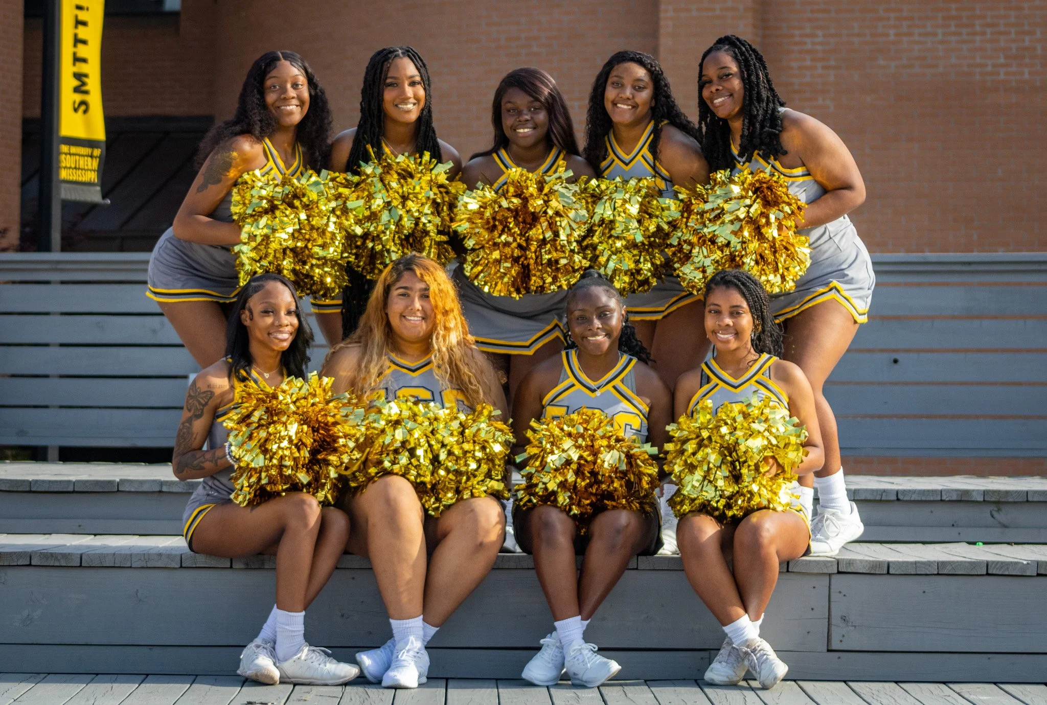 A group of ten young women in cheerleading uniforms and holding gold pom-poms posing on outdoor bleachers.