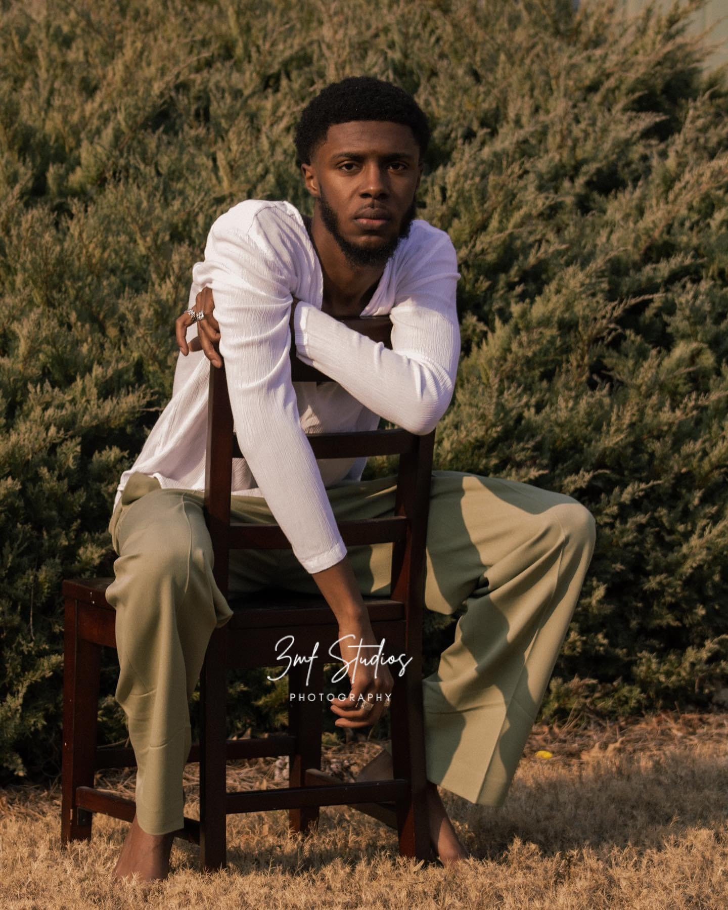A man in a white shirt and beige pants sitting on a wooden chair outdoors in front of green bushes, with a serious expression, during sunset.