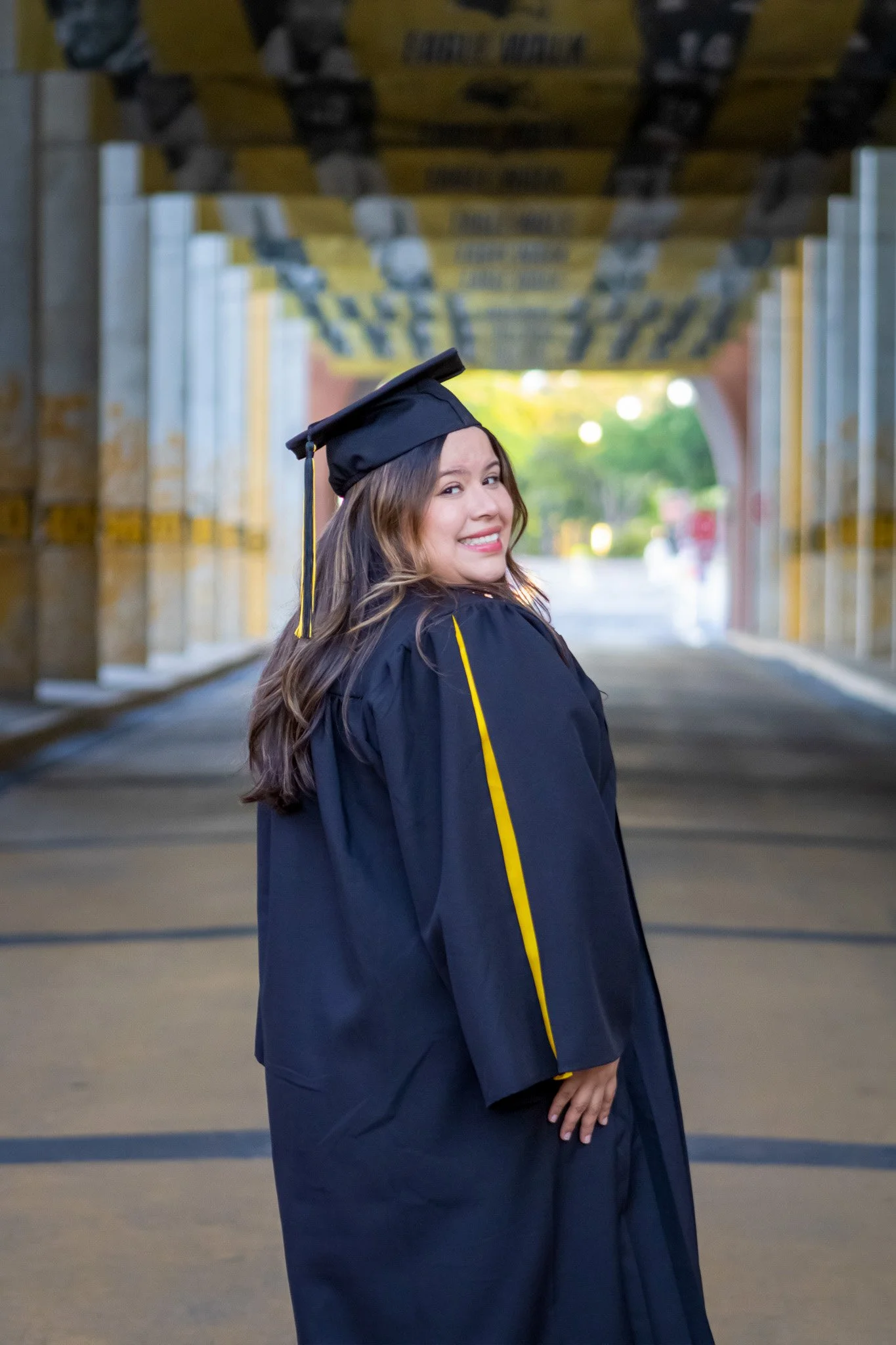 Woman in graduation cap and gown smiling at camera under a bridge.