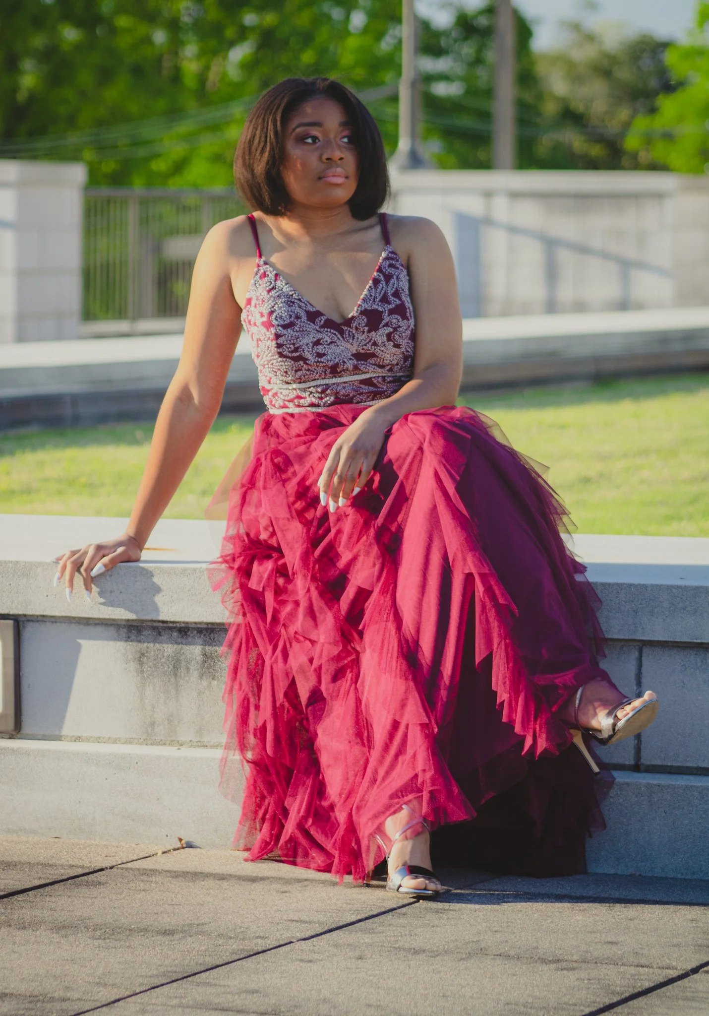 A woman in a red and white lace top and a flowing red tulle skirt sitting on a concrete ledge outdoors on a sunny day.