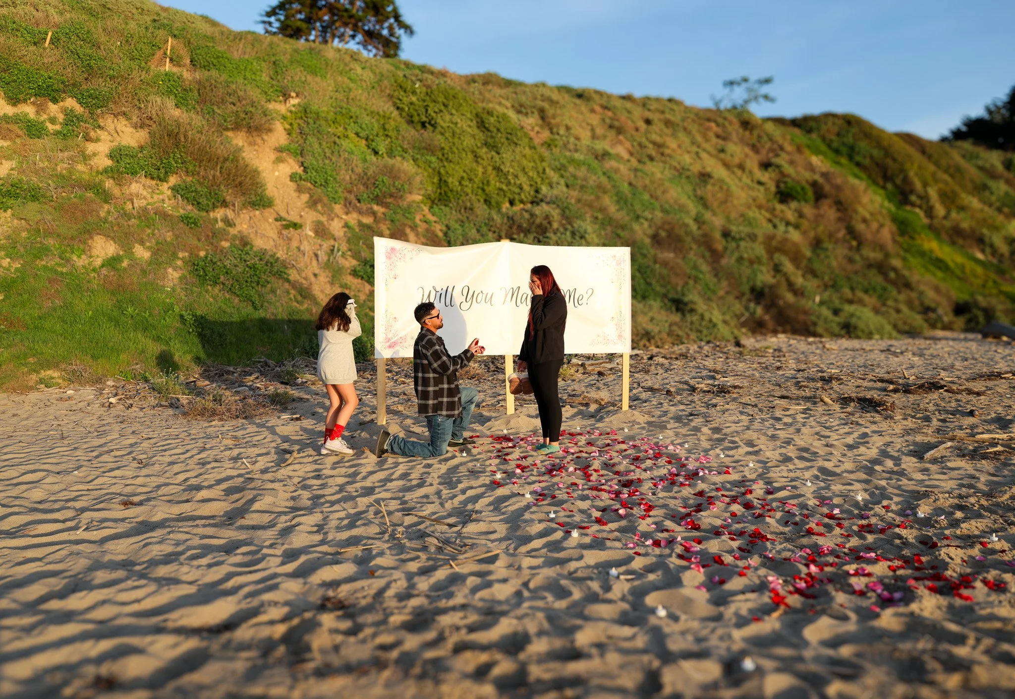 A man proposing to a woman on a beach with rose petals arranged in a heart shape and a sign that reads "Will You Marry Me?". A girl stands nearby watching the scene. The background features green hills under a clear blue sky.