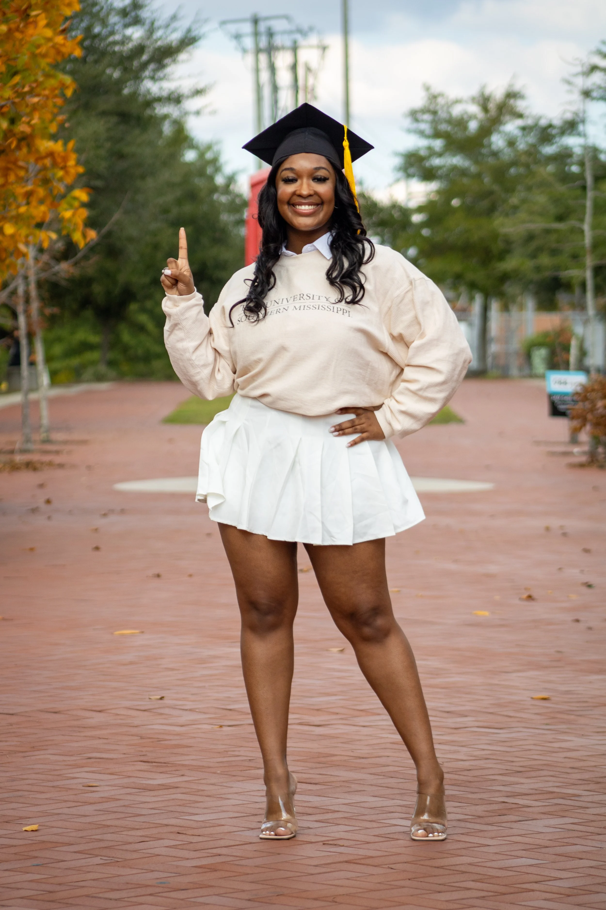 A woman in a graduation cap and gown on campus, smiling and pointing upward.