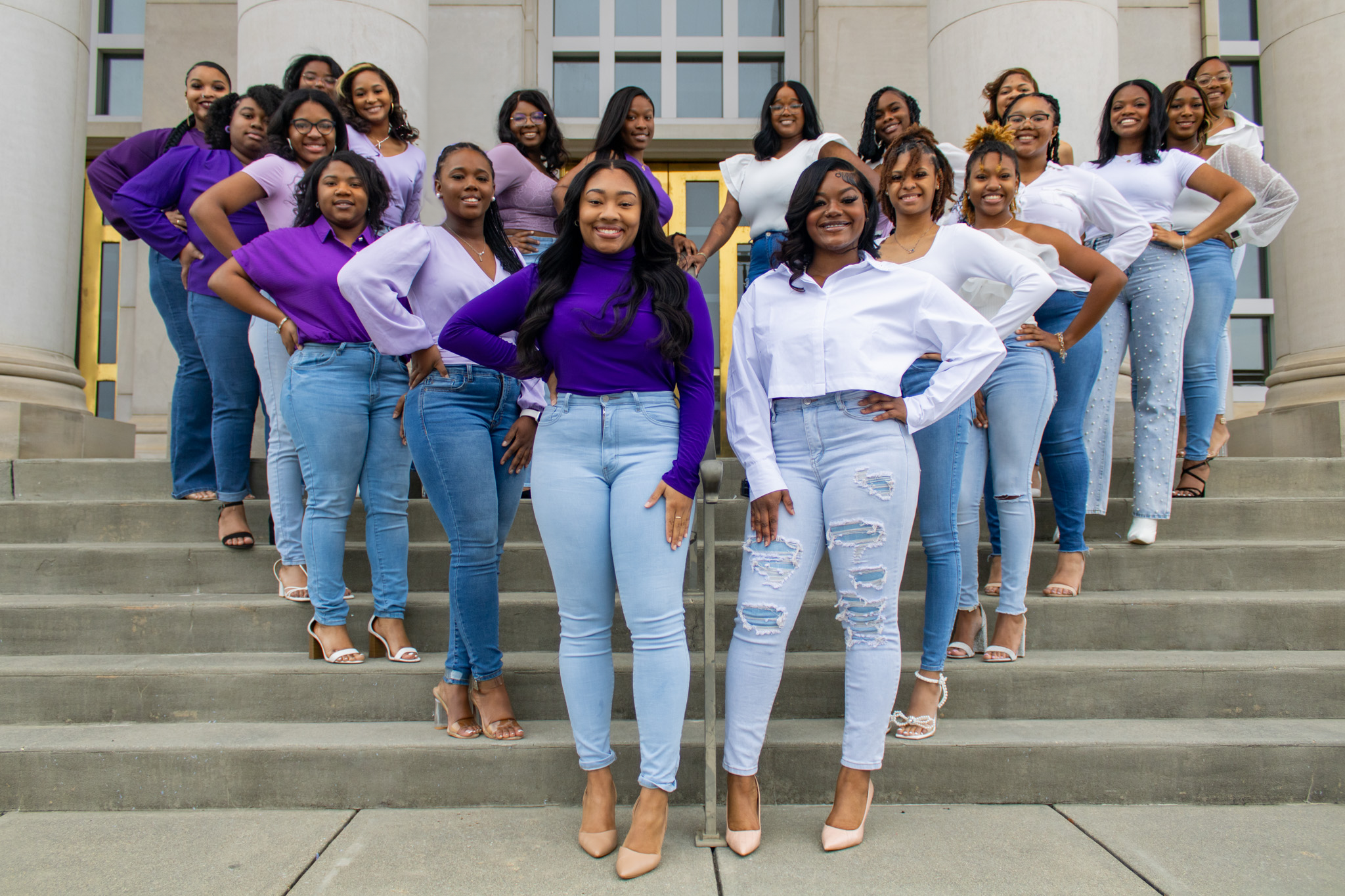 A large group of women standing on the steps of a government building or courthouse, wearing coordinated casual outfits in shades of purple, white, and blue, smiling and posing for the photo.