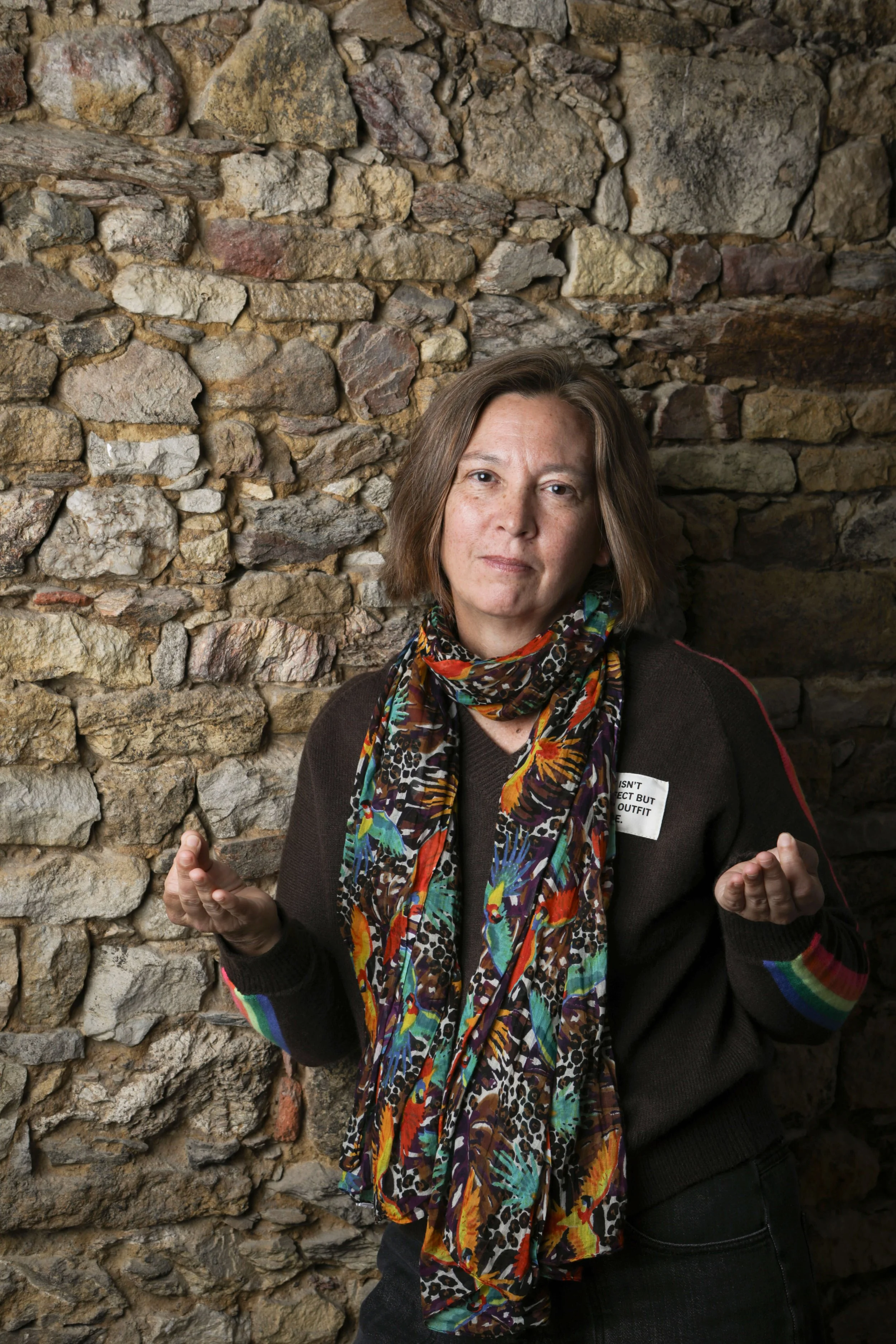 Une femme debout contre un mur en pierre, portant un pull sombre et une écharpe colorée avec un motif animal, faisant un geste avec ses mains et regardant vers l'avant.