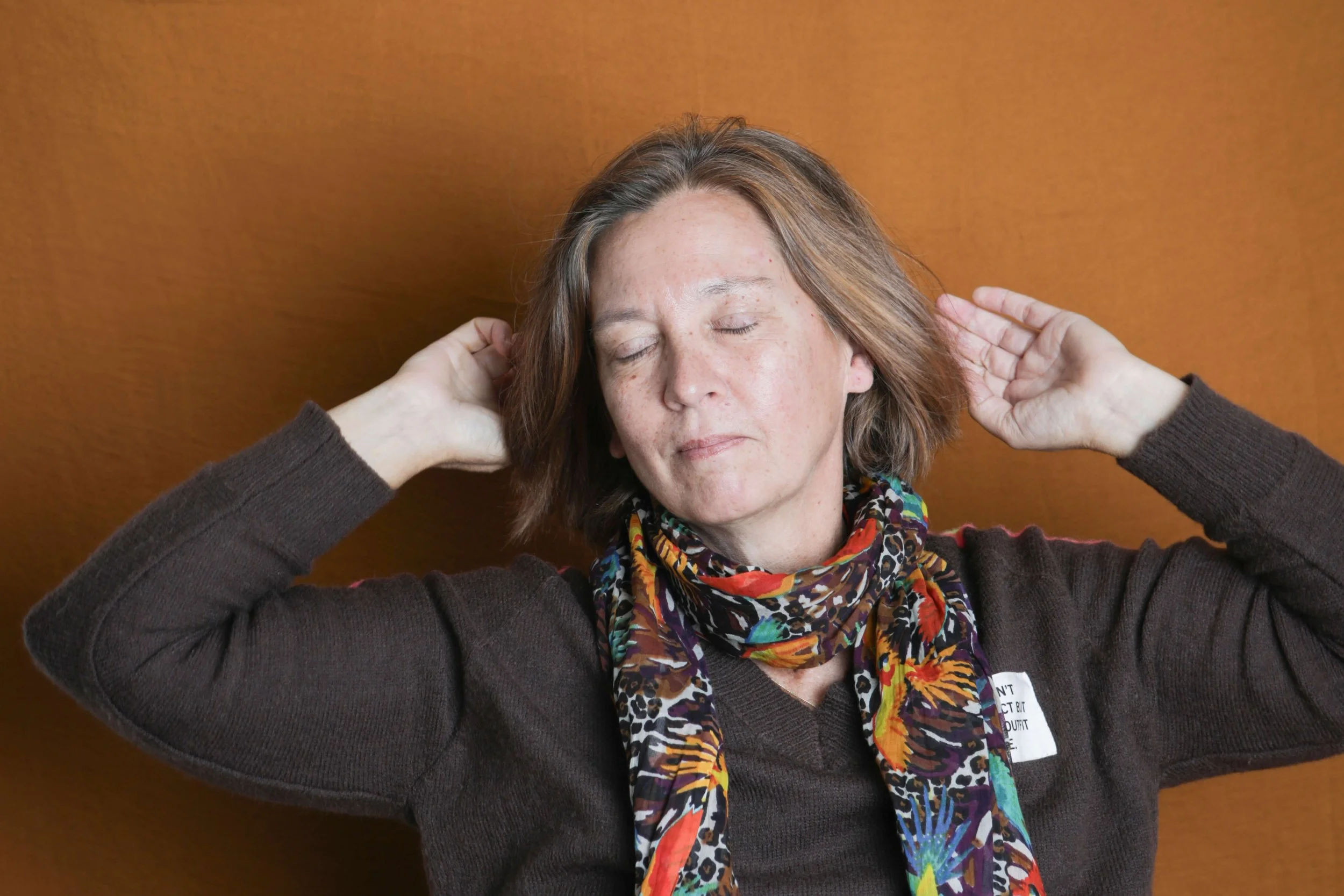 Une femme aux cheveux courts et bruns, les yeux fermés, souriant légèrement, portant un foulard coloré et un pull marron, se détend contre un mur orange.