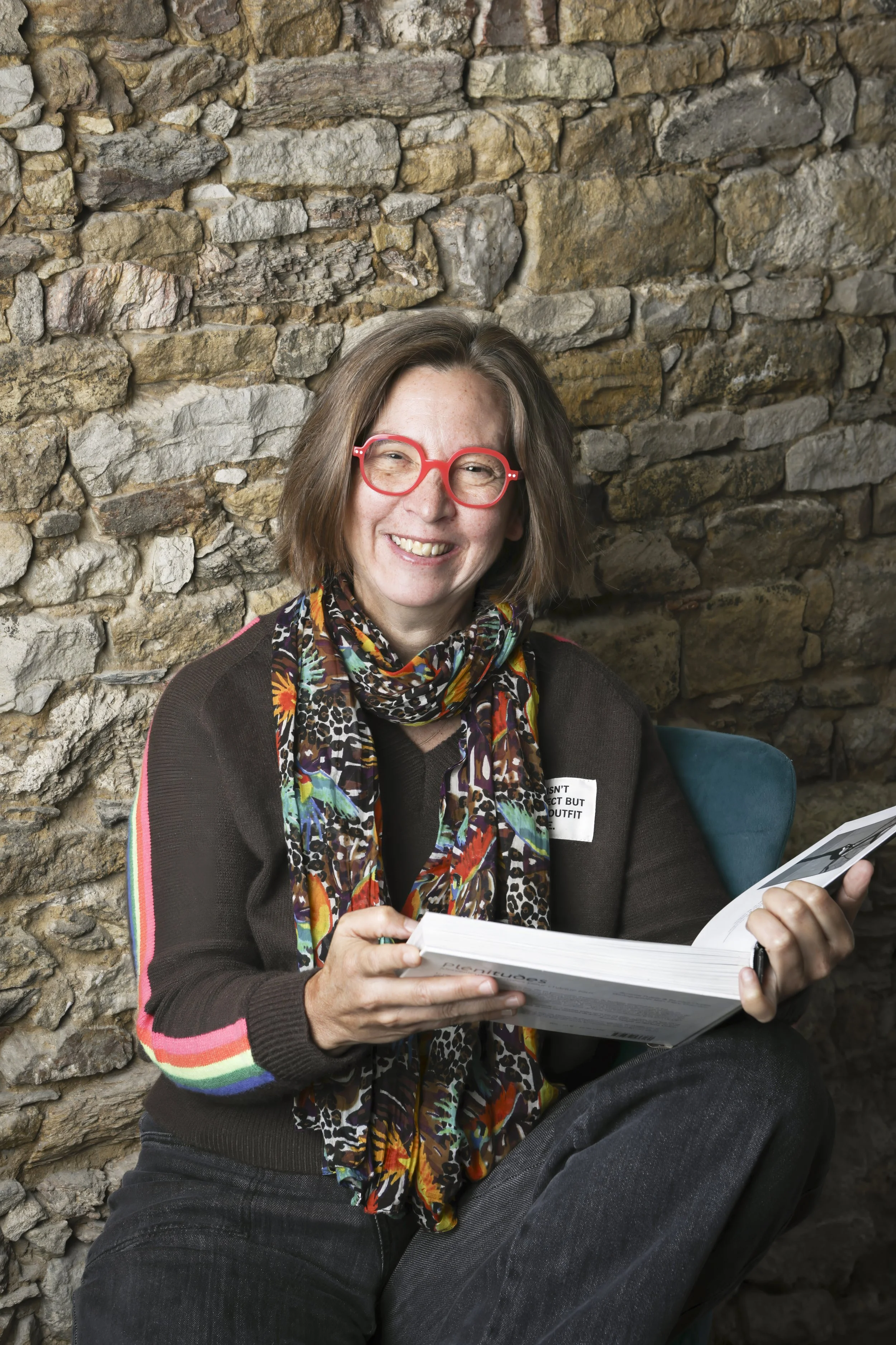 Femme souriante avec des lunettes rouges, portant un foulard coloré, assise contre un mur en pierre, lisant un livre.