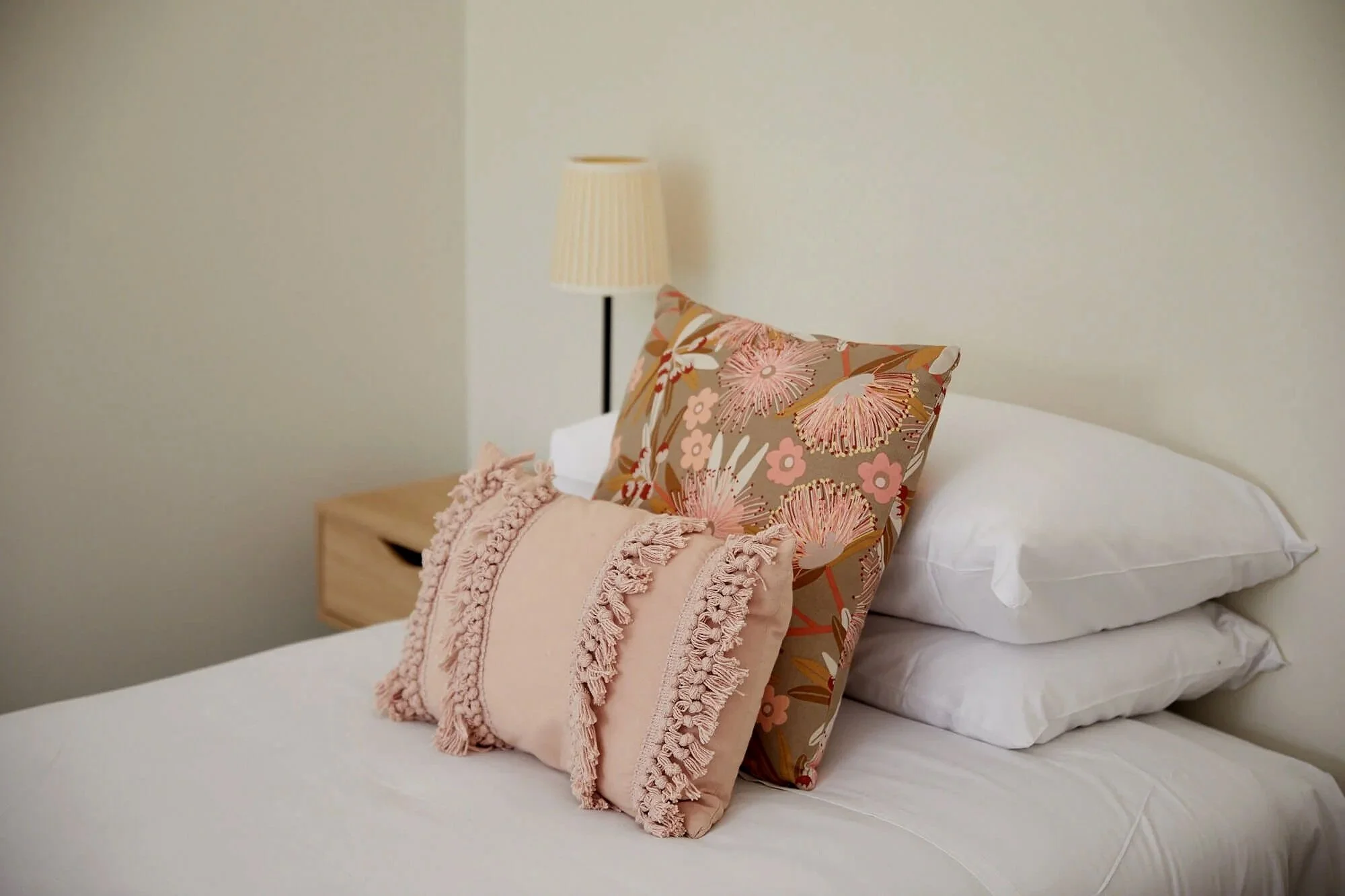Bed with two decorative pillows, one with tassels and another with a floral pattern, on top of white pillows and sheets; a nightstand and lamp in the background.