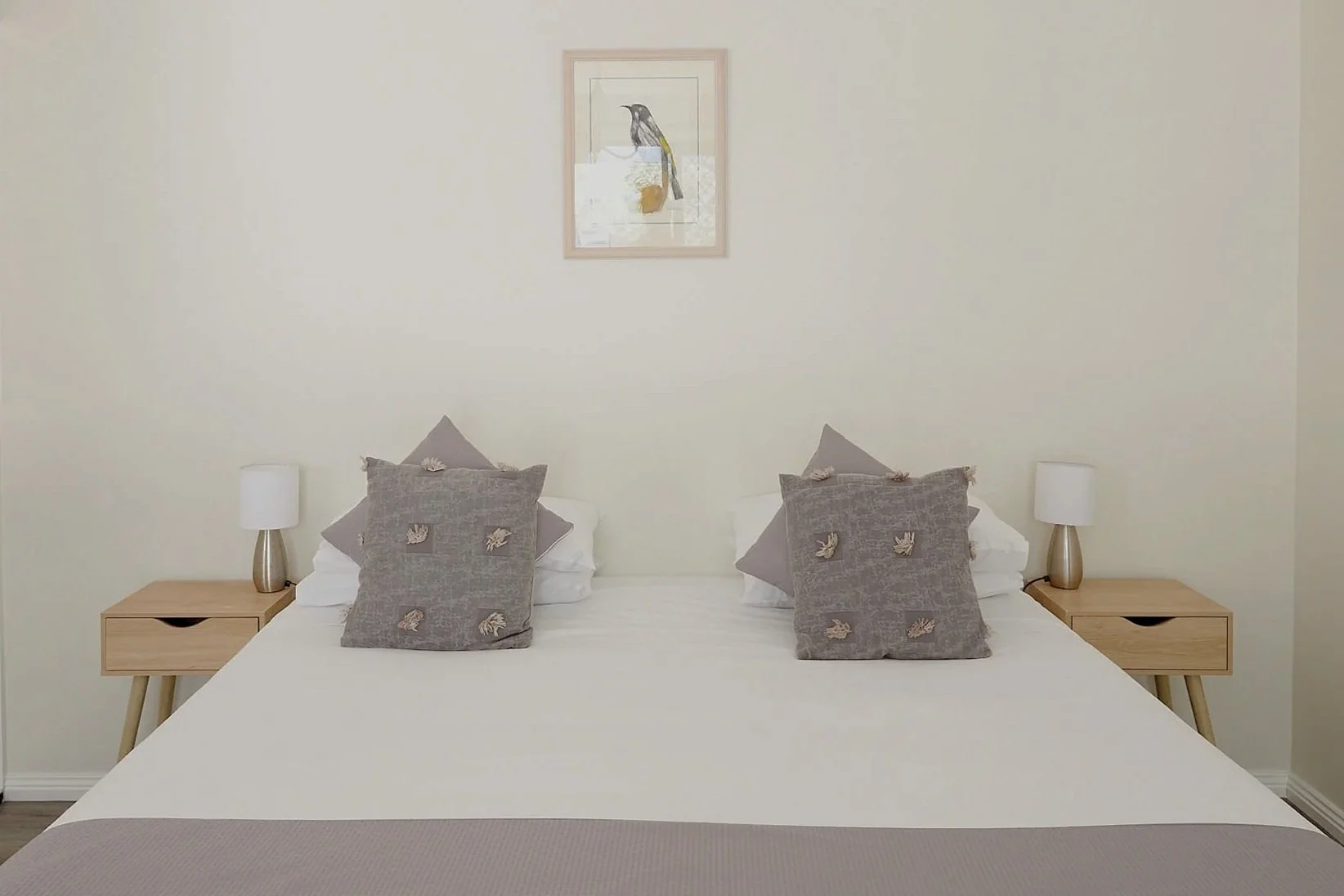 Minimalist bedroom with neatly made bed, two gray decorative pillows, wooden nightstands, small lamps, and framed bird art on white wall.