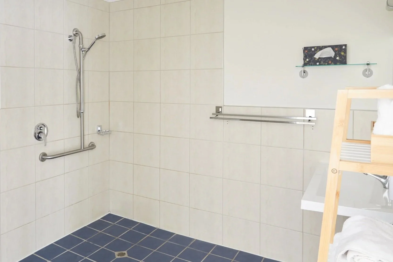 Accessible bathroom with beige tiled walls, blue tiled floor, grab bars, and handheld showerhead. Wooden shelf with towels and a sink are visible.