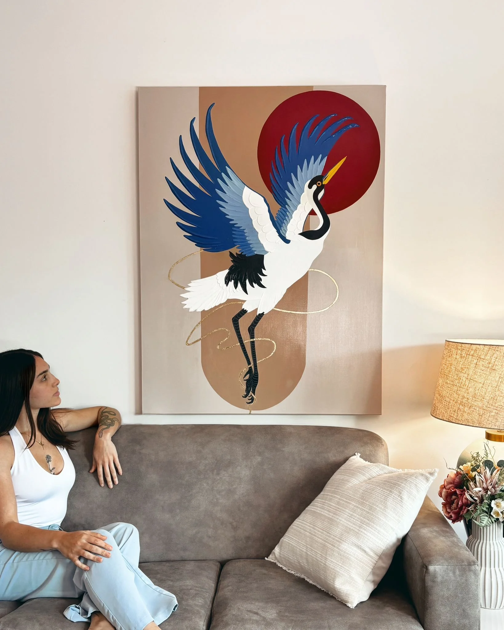A woman sits on a gray sofa beside a beige pillow, looking at a large modern painting of a crane with blue and black feathers against a beige and red background, with a lamp and floral arrangement nearby.