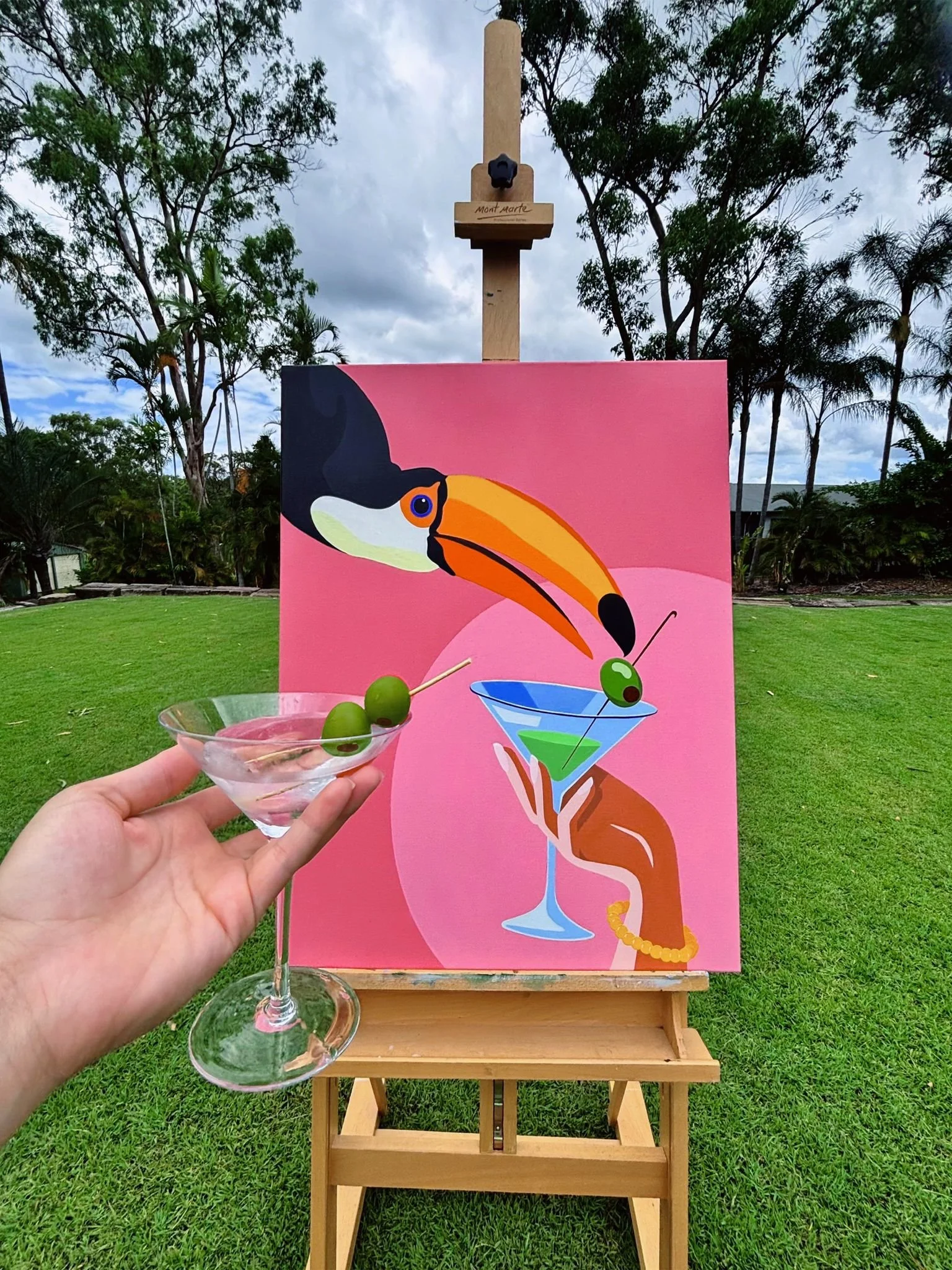 A hand holding a martini glass with two green olives and a toothpick, in front of a colorful painting of a toucan and a martini glass on a stand, outdoors on a grassy area with trees and cloudy sky in the background.