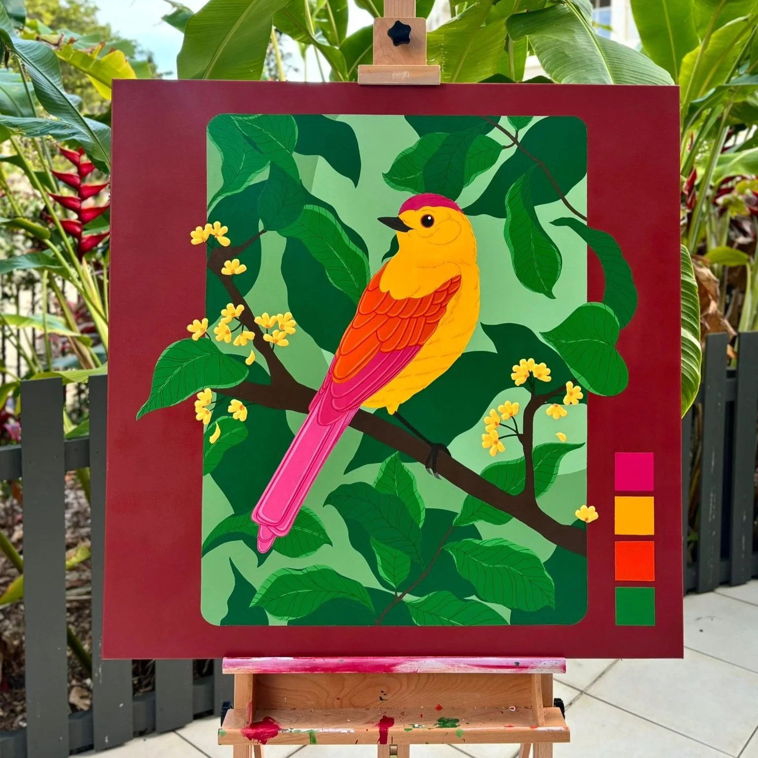 Colorful painting of a yellow and pink bird perched on a branch among green leaves and yellow flowers, framed in a red border, outdoors with plants in the background.