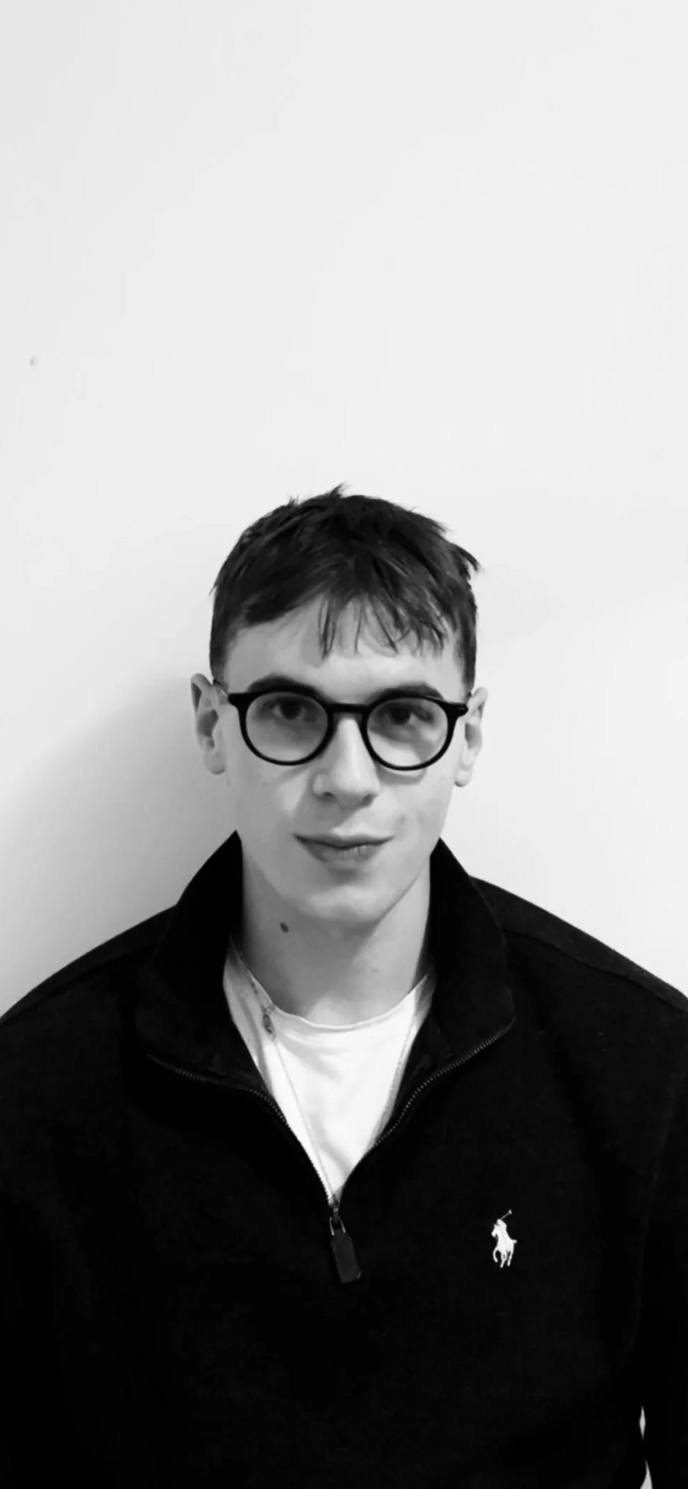 Black and white portrait of a young man with short dark hair and glasses, wearing a dark zipped jacket with a Polo Ralph Lauren logo, standing against a plain white background.