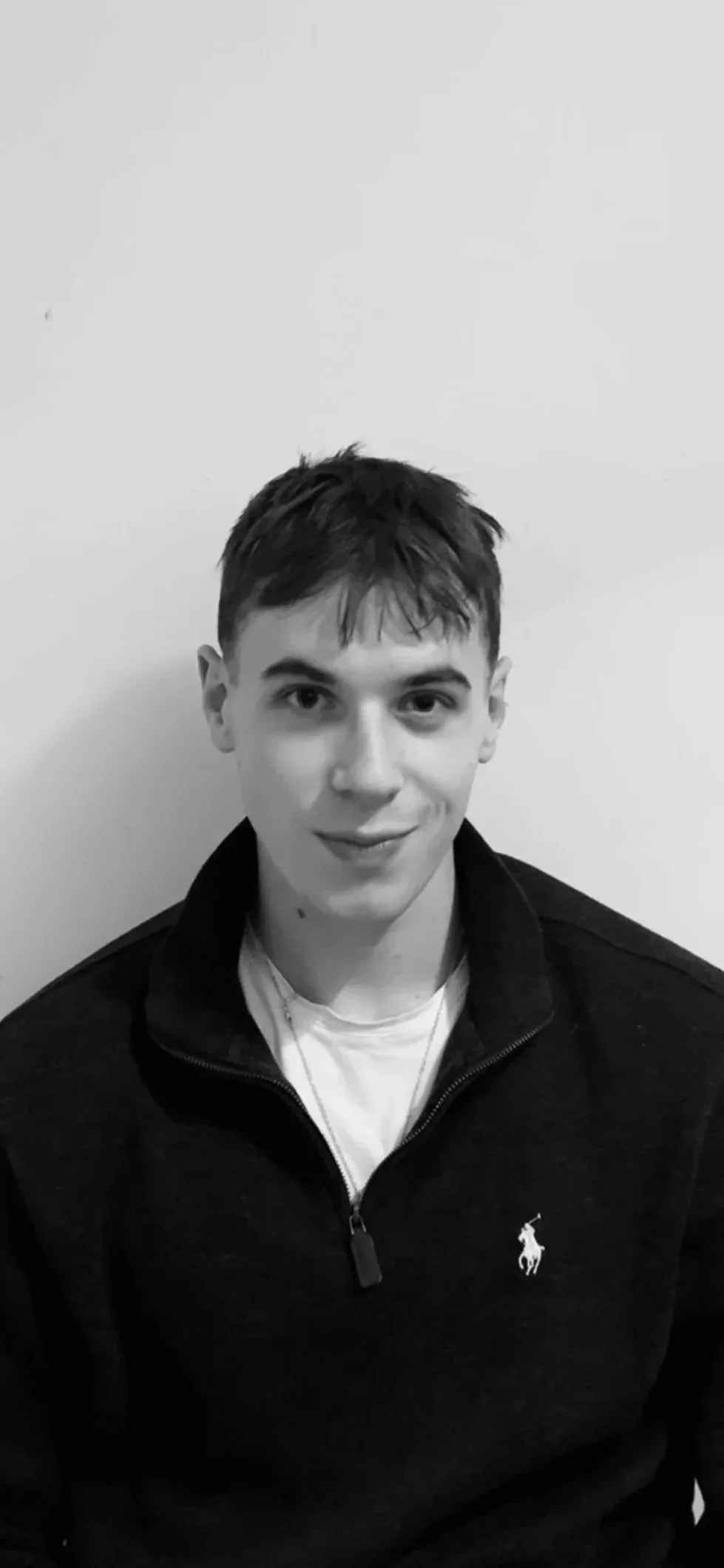 Black and white photo of a young man with short hair, smiling, wearing a dark zip-up jacket with a Polo Ralph Lauren logo, standing against a plain wall.