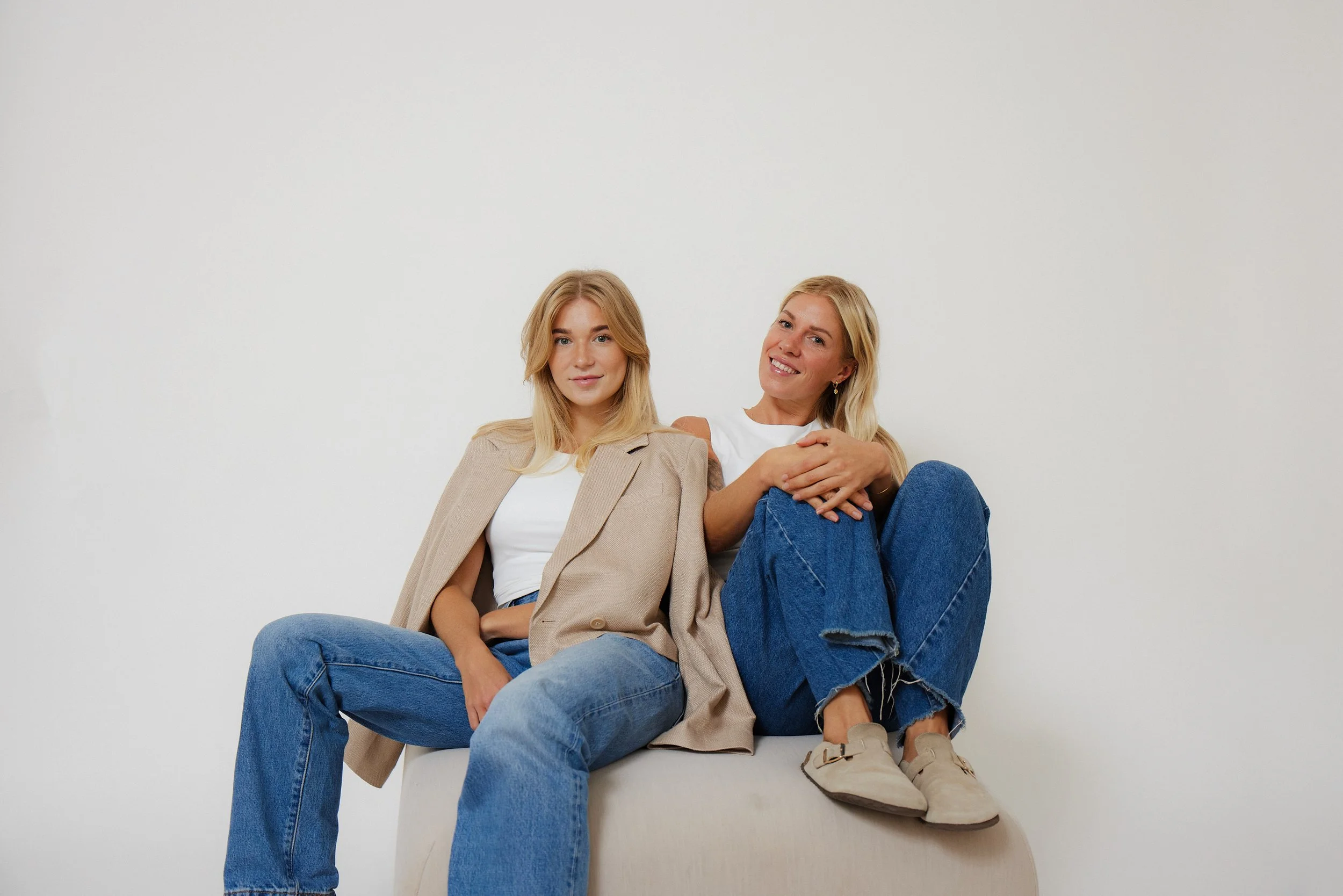 Two women sitting on a white ottoman against a plain white wall, one wearing a beige blazer and jeans, the other wearing a white t-shirt and blue jeans, both smiling at the camera.