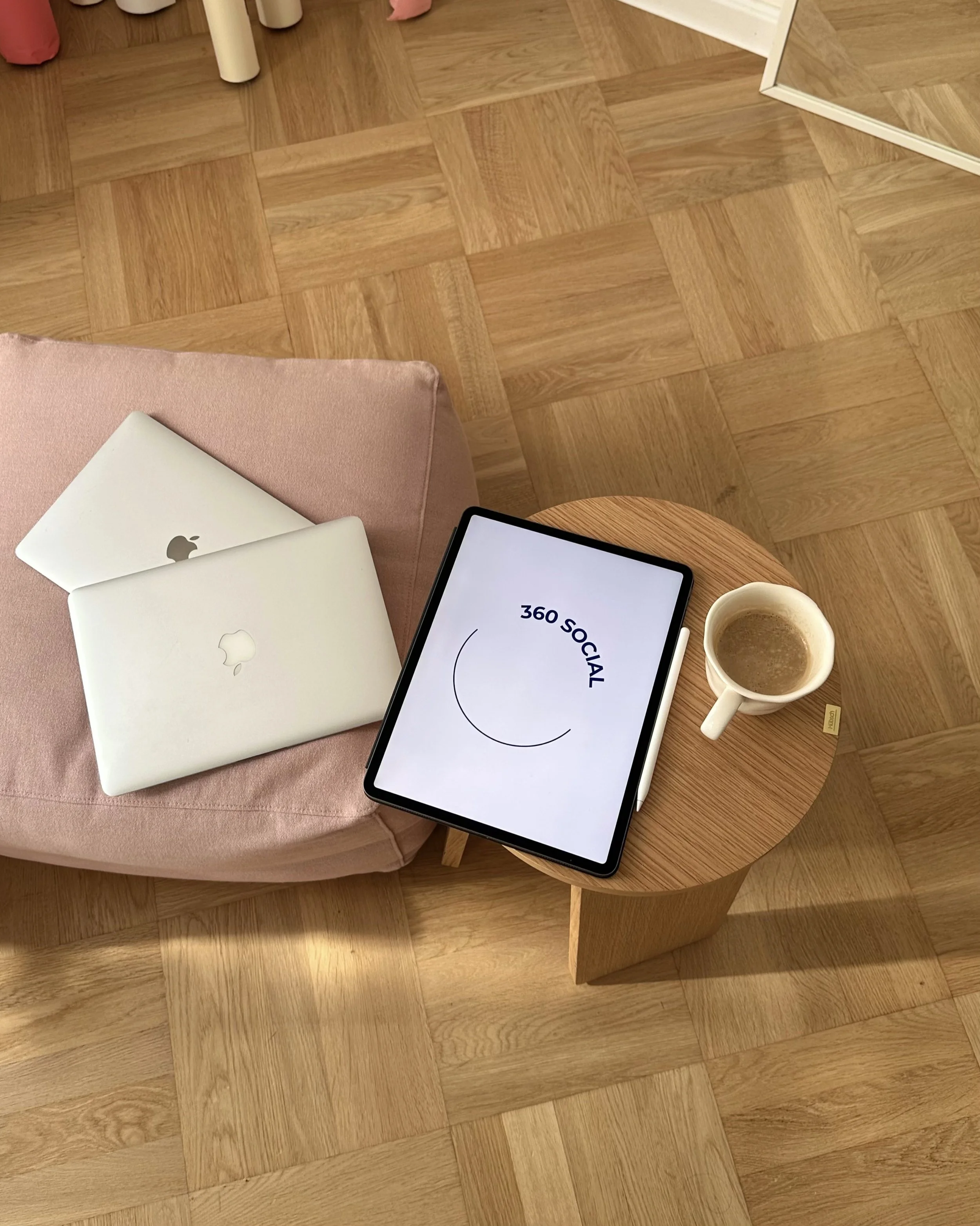 A pink cushioned seat with two closed Apple MacBook laptops on it, a small round wooden side table with a tablet displaying '360 SOCIAL' and a cup of coffee, and a wooden parquet floor.