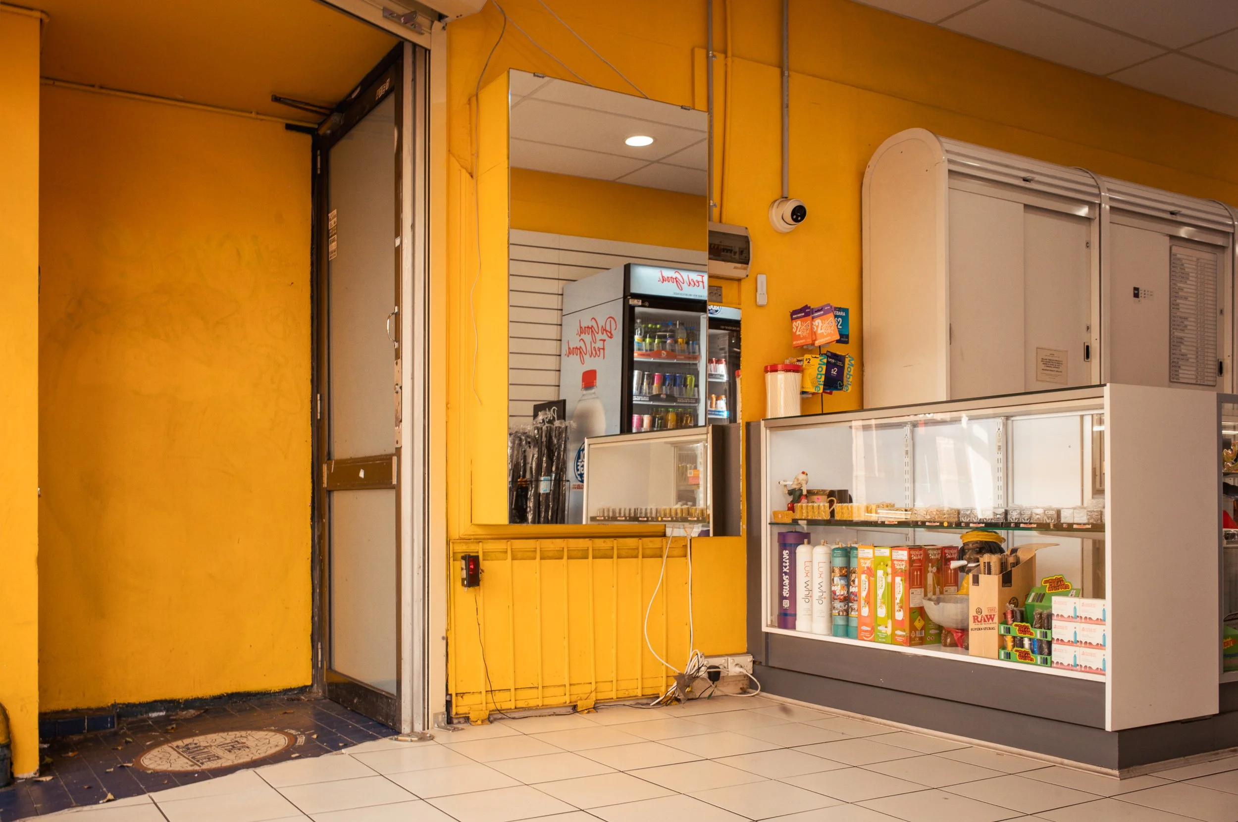Interior of a convenience store with yellow painted walls, a glass display case filled with snacks and products, a small refrigerator with drinks, and a partially open door leading outside.