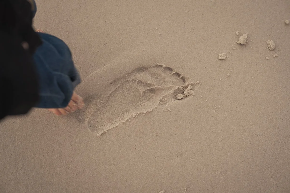 A person standing on a sandy beach, digging a hole in the sand with their bare feet and toes.