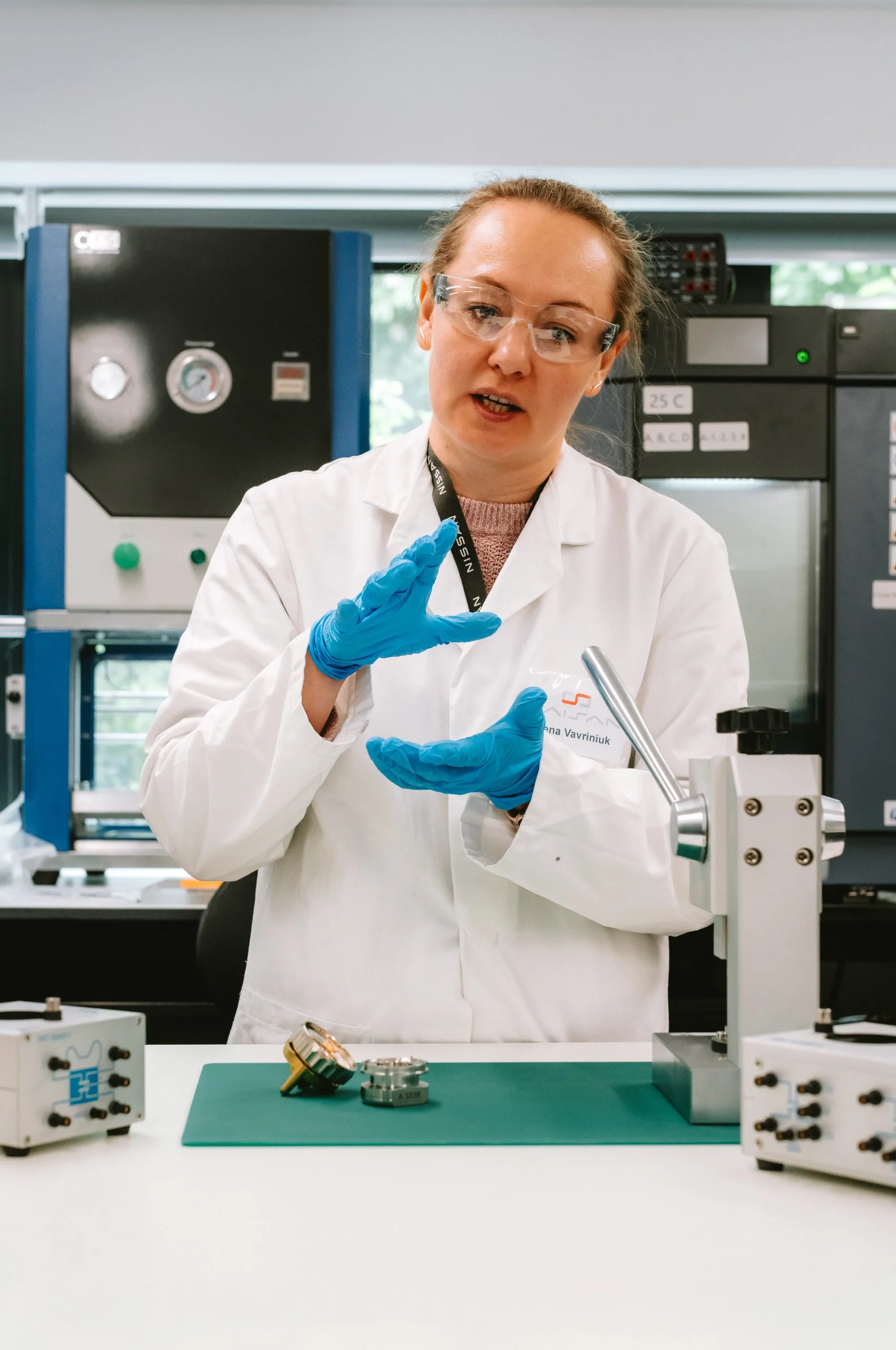 A woman wearing safety glasses, blue gloves, and a white lab coat is standing in a laboratory, speaking and gesturing with her hands, with scientific equipment and machinery in the background.