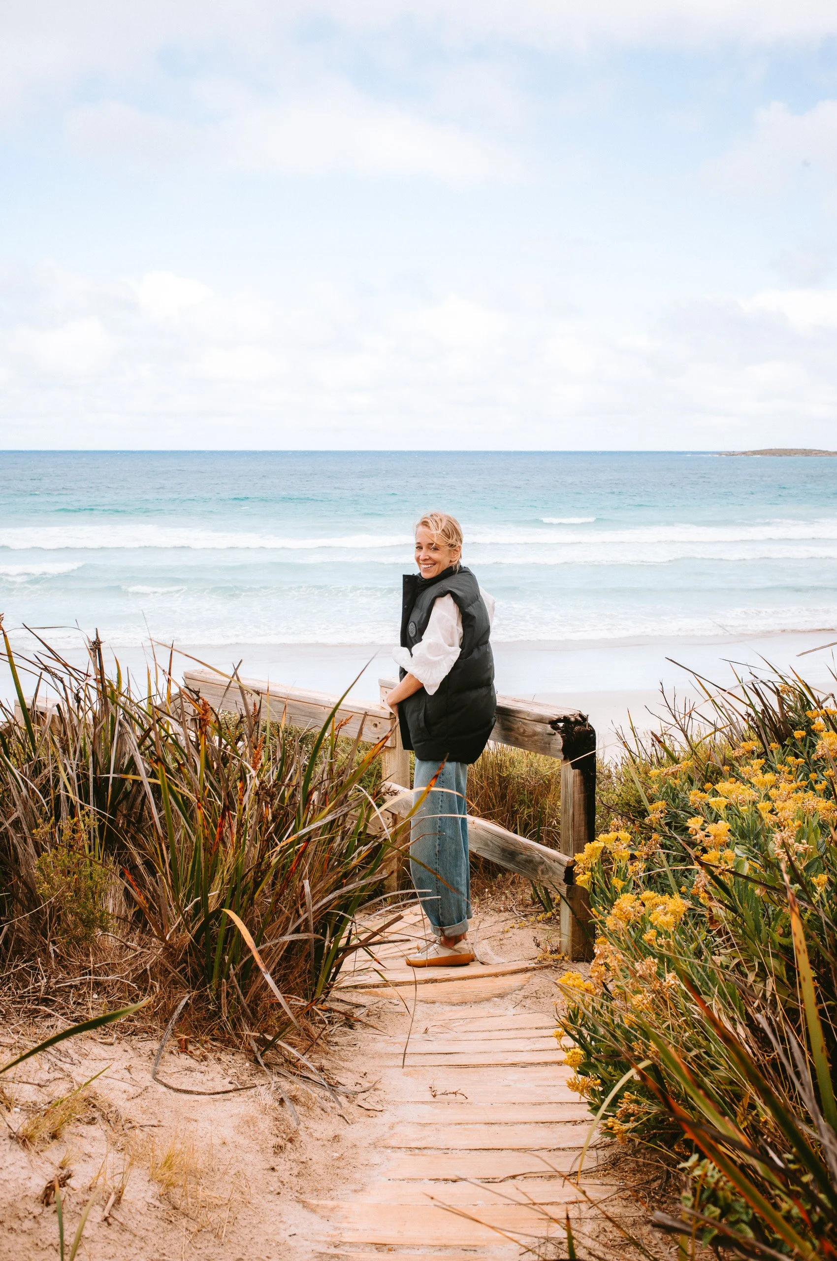 A woman standing on a wooden pathway leading to a beach, smiling at the camera, with dry grass and yellow flowers on either side and the ocean in the background.