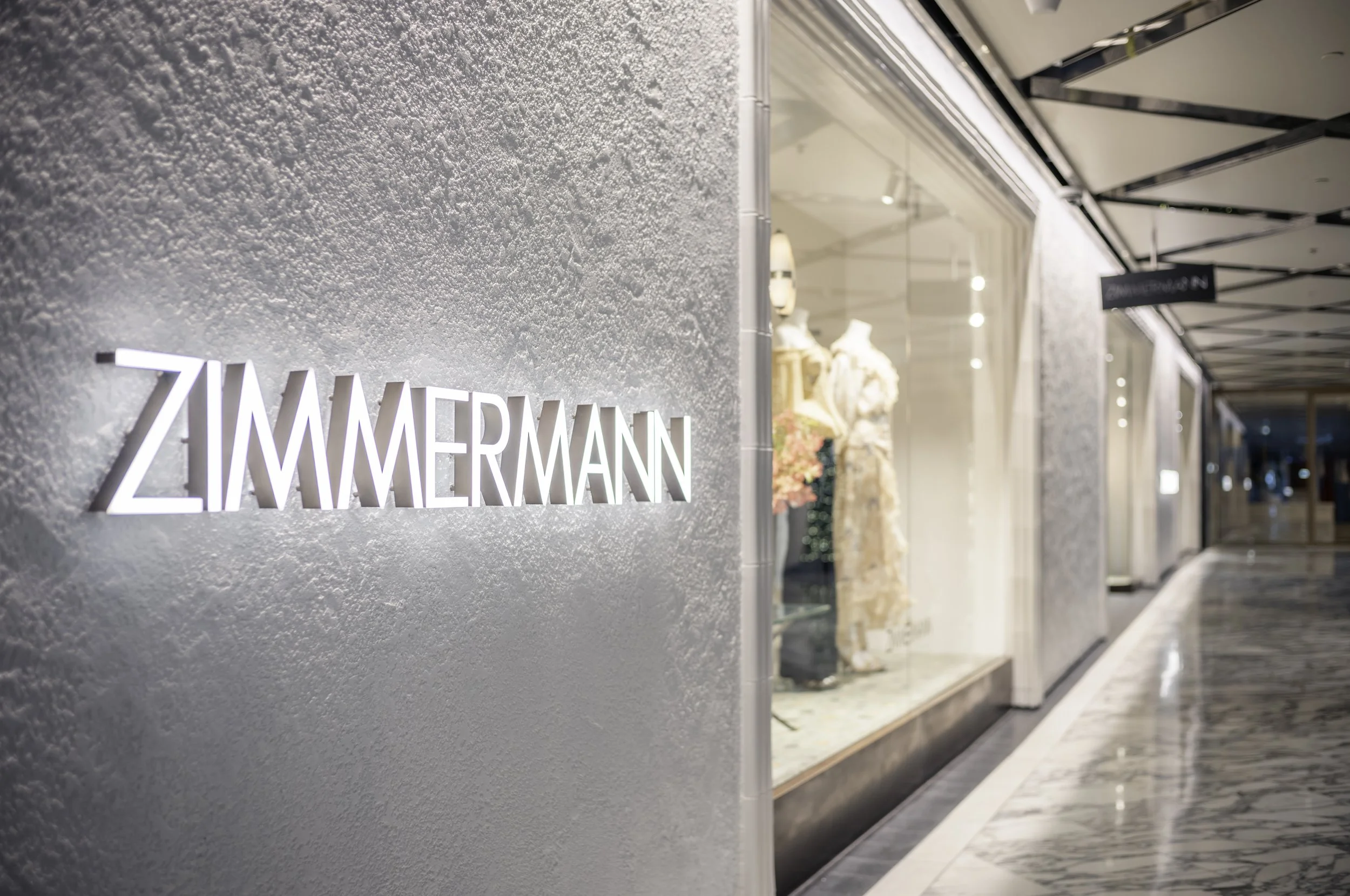ZIMMERMANN store front with illuminated sign, mannequins in dresses displayed inside, marble flooring in a mall corridor.