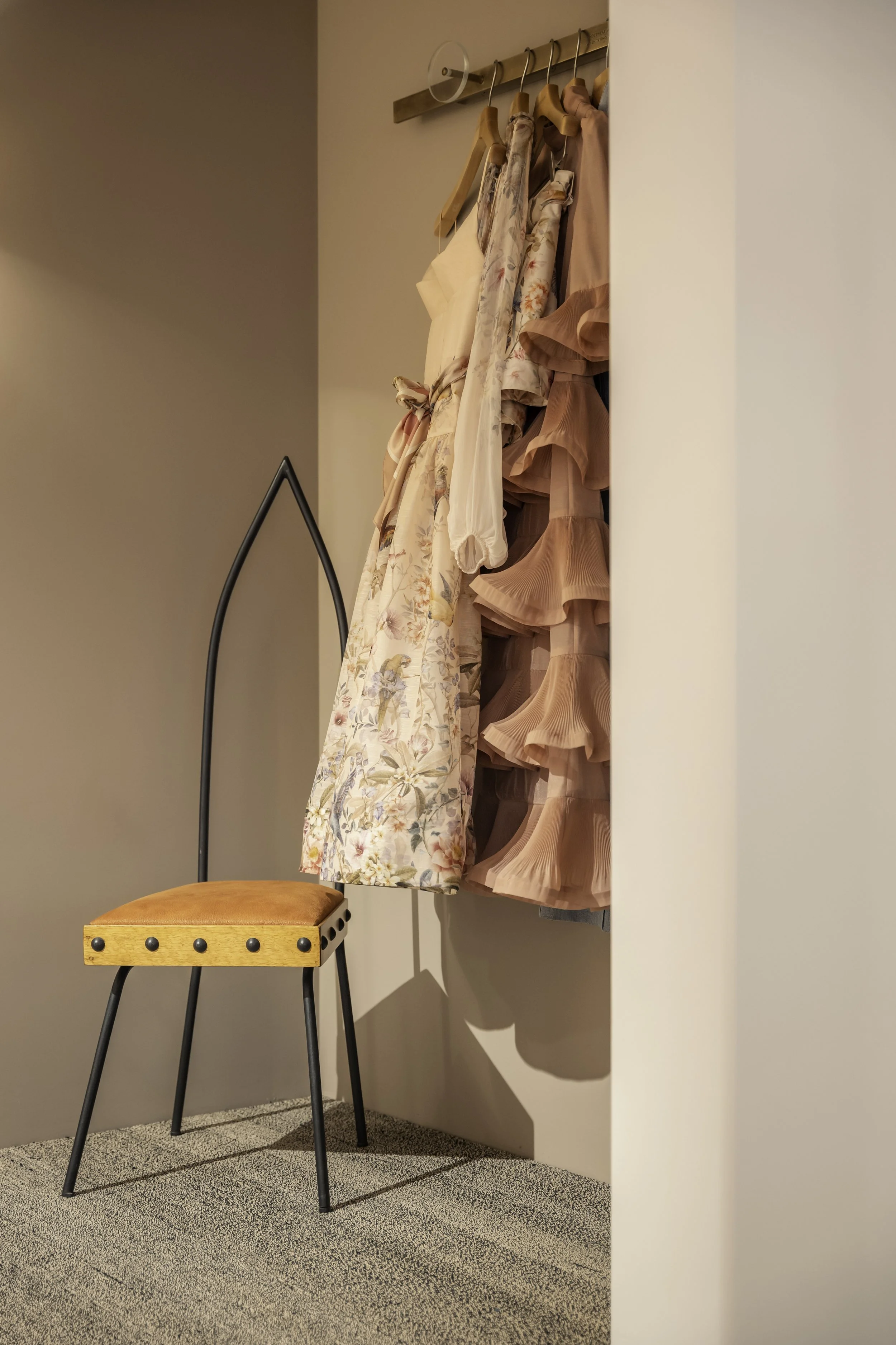 A corner of a room with a small wooden and metal chair with orange cushion, and a wall-mounted rack with hanging dresses and skirts in pastel pink, beige, and floral patterns.