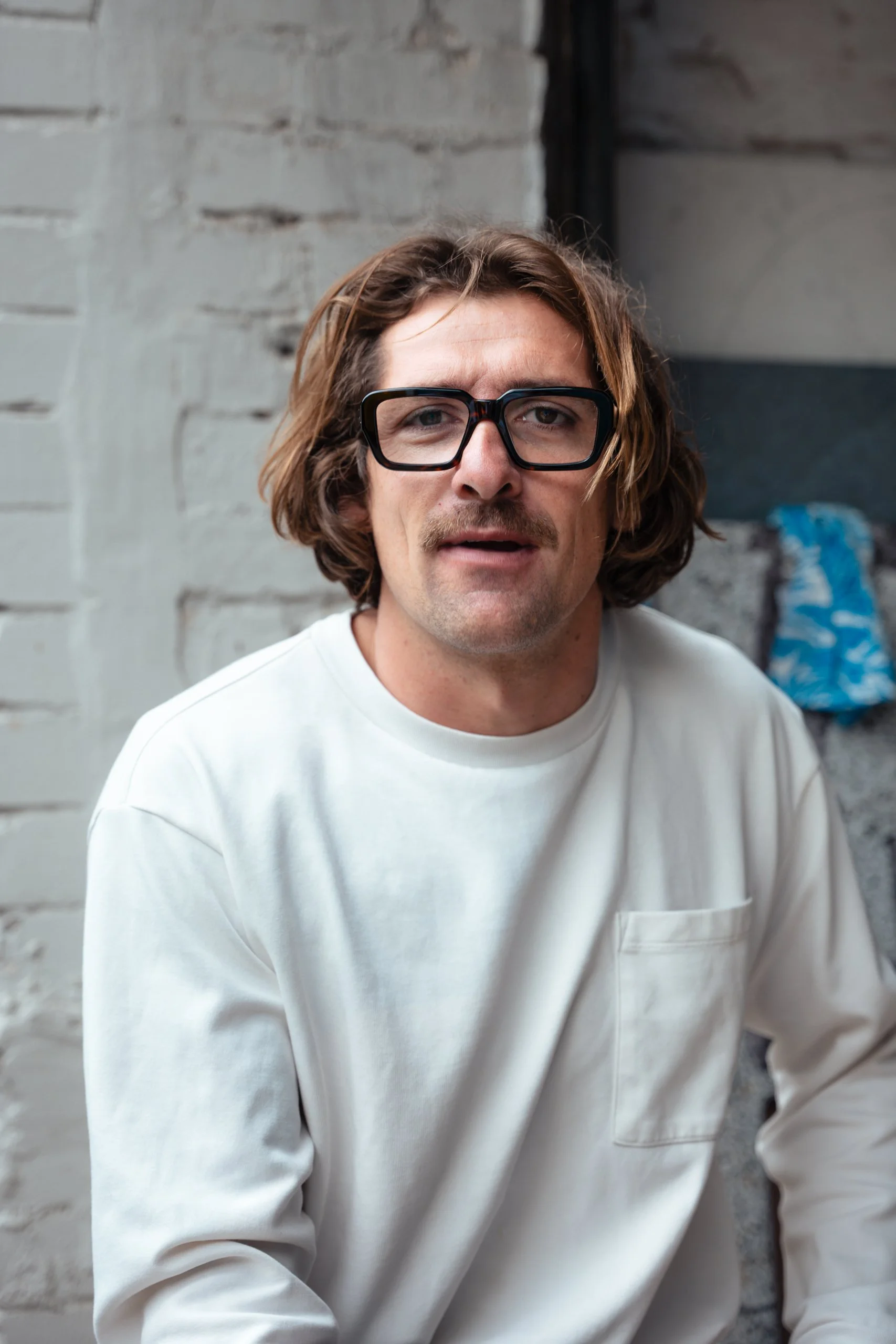 A man with medium-length wavy brown hair, wearing large black glasses and a white long-sleeve shirt, is sitting against a brick wall with a slightly puzzled or questioning expression.