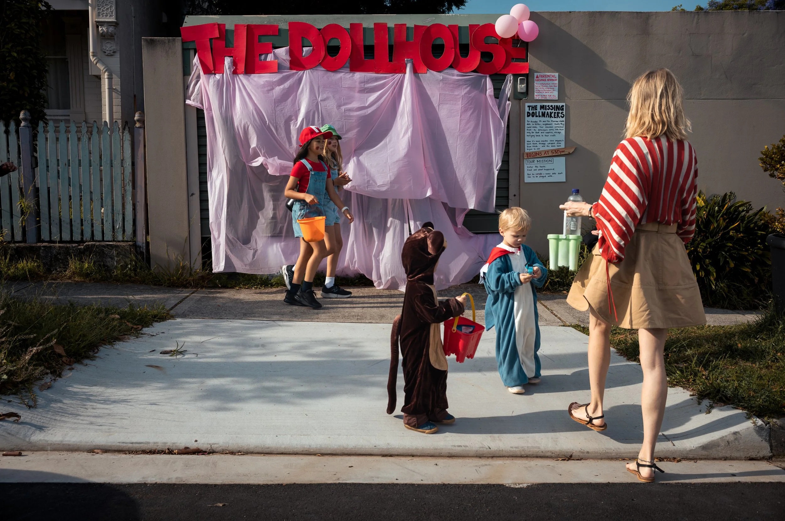 Children and a woman dressed in costumes trick-or-treat during Halloween night. A house decorated with pink fabric and balloons, with a large sign reading 'The Dollhouse.'