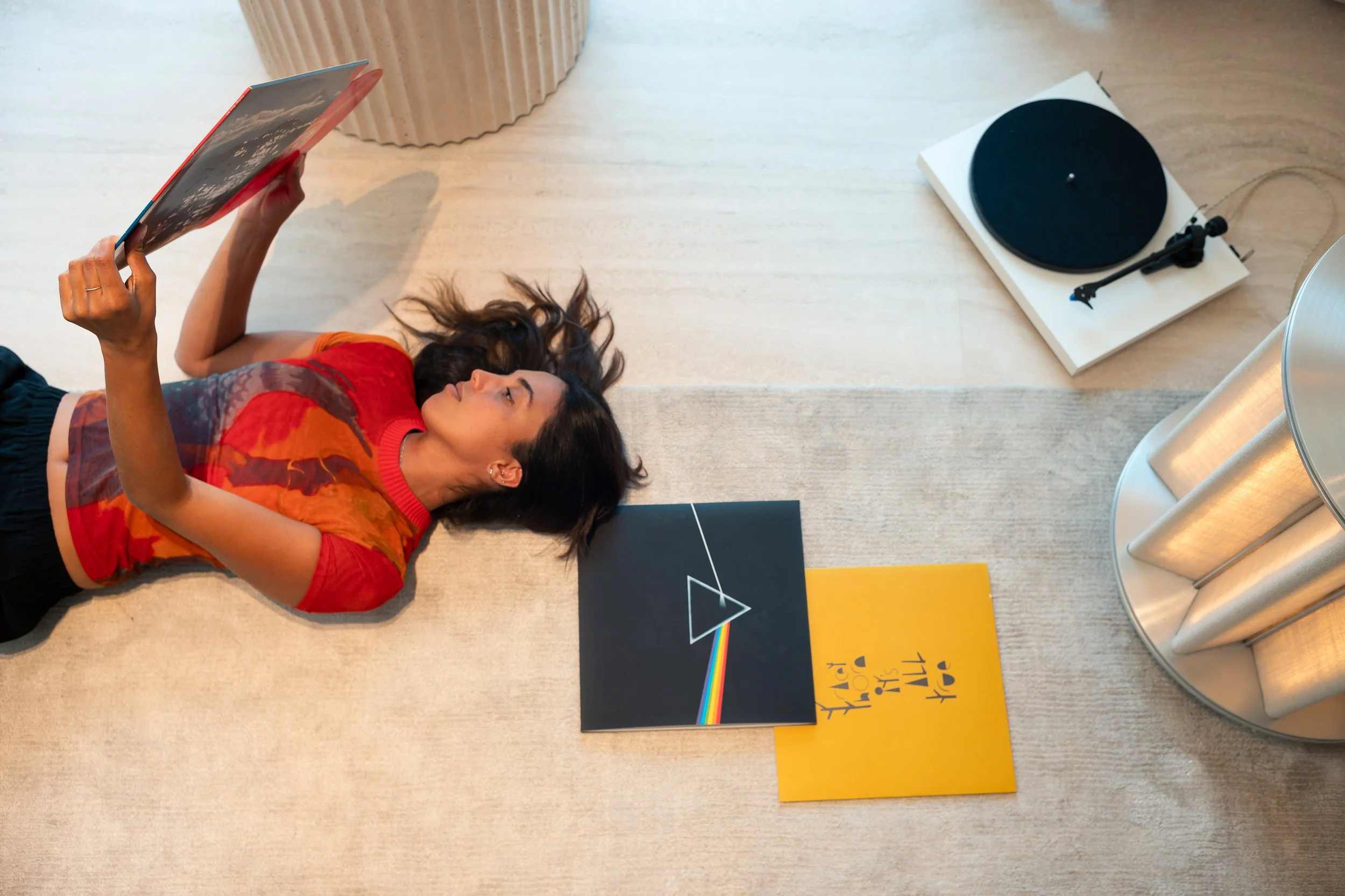 Young woman lying on floor, reading a record album with Pink Floyd's "The Dark Side of the Moon" on the cover, which features a prism and rainbow. There are additional record albums nearby and a turntable to the side.