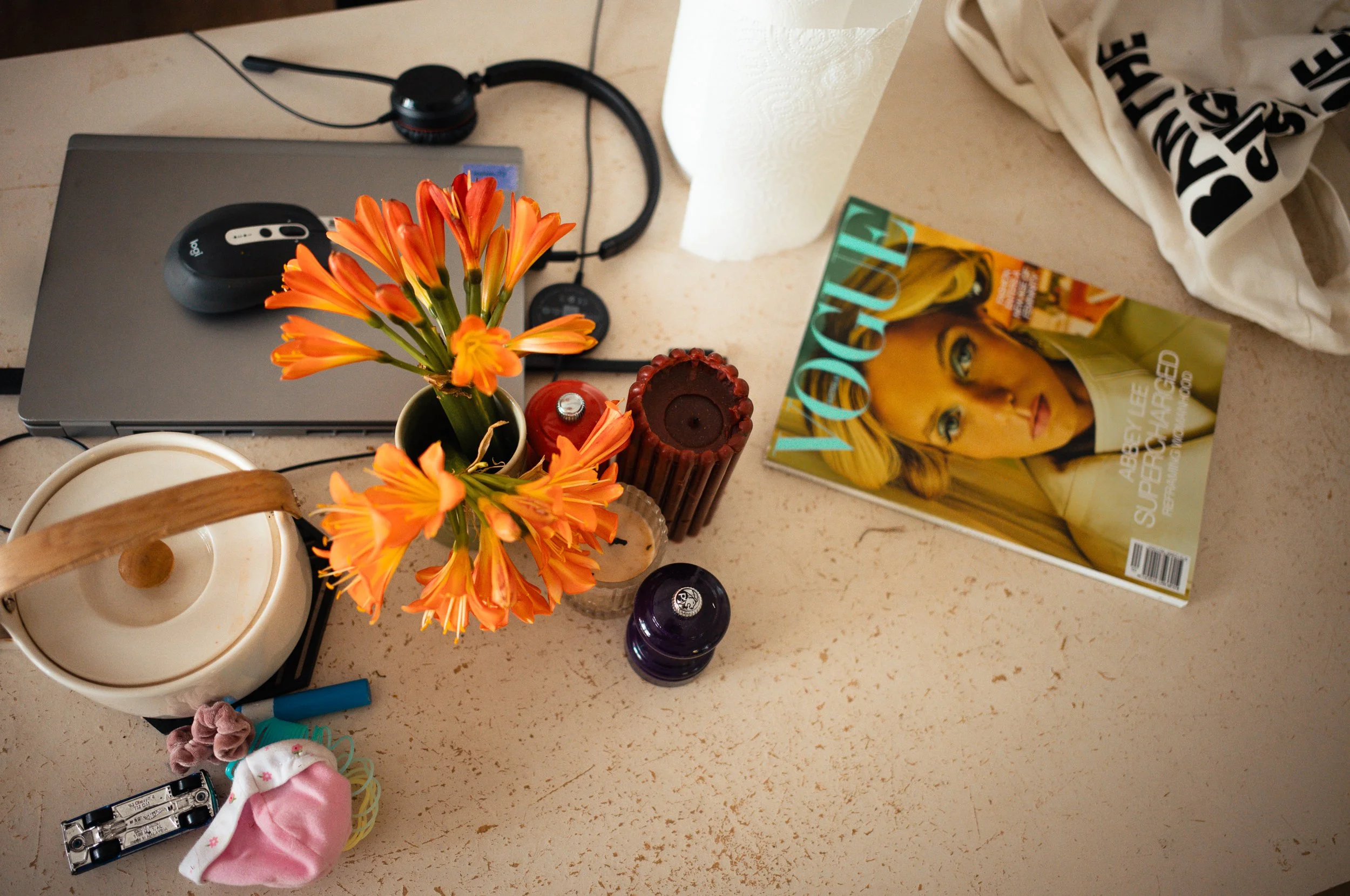 A beige countertop with orange flowers in a small black pot, a magazine titled 'VOGUE' featuring a blonde woman on the cover, a white paper towel roll, a beige cloth bag, a laptop with a computer mouse, a red candle, a purple bottle, a teapot, a smal