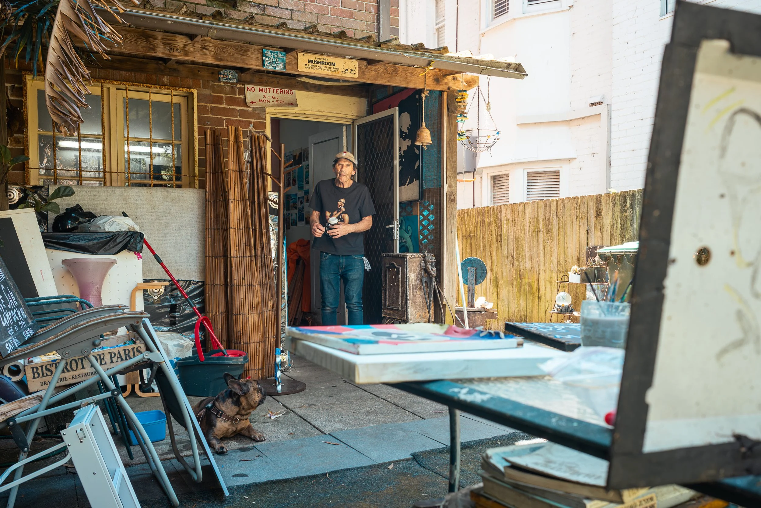 A man standing in the doorway of a cluttered backyard or garage, holding a mug, with a small dog sitting on the ground nearby, surrounded by various outdoor and painting supplies.