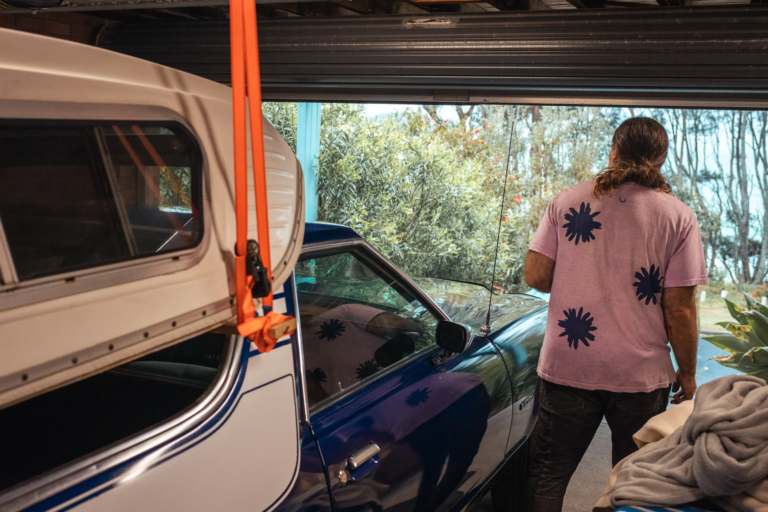 A man with long hair, wearing a pink shirt with black floral prints, stands in a garage next to a black vintage car. The garage door is open, revealing greenery and trees outside.