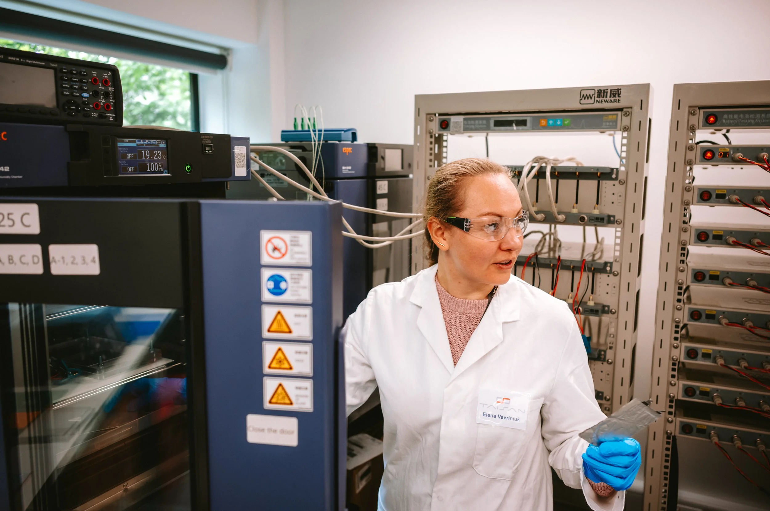 A woman in a white lab coat and safety glasses working with laboratory equipment, holding a small object in a plastic bag, in a laboratory with electrical panels and scientific instruments.