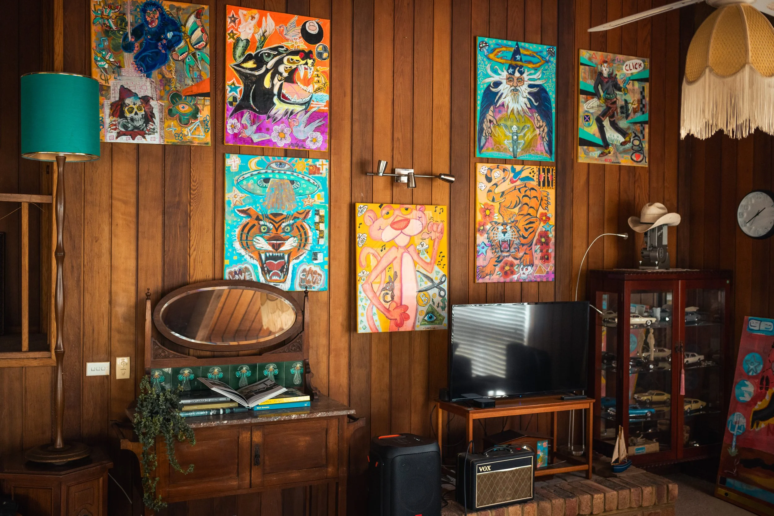 A wooden-paneled living room wall decorated with eight colorful, contemporary paintings of various animals and fantasy themes. There is a teal floor lamp, a vintage mirror, a small bookshelf, a flat-screen TV, and a display cabinet filled with model 