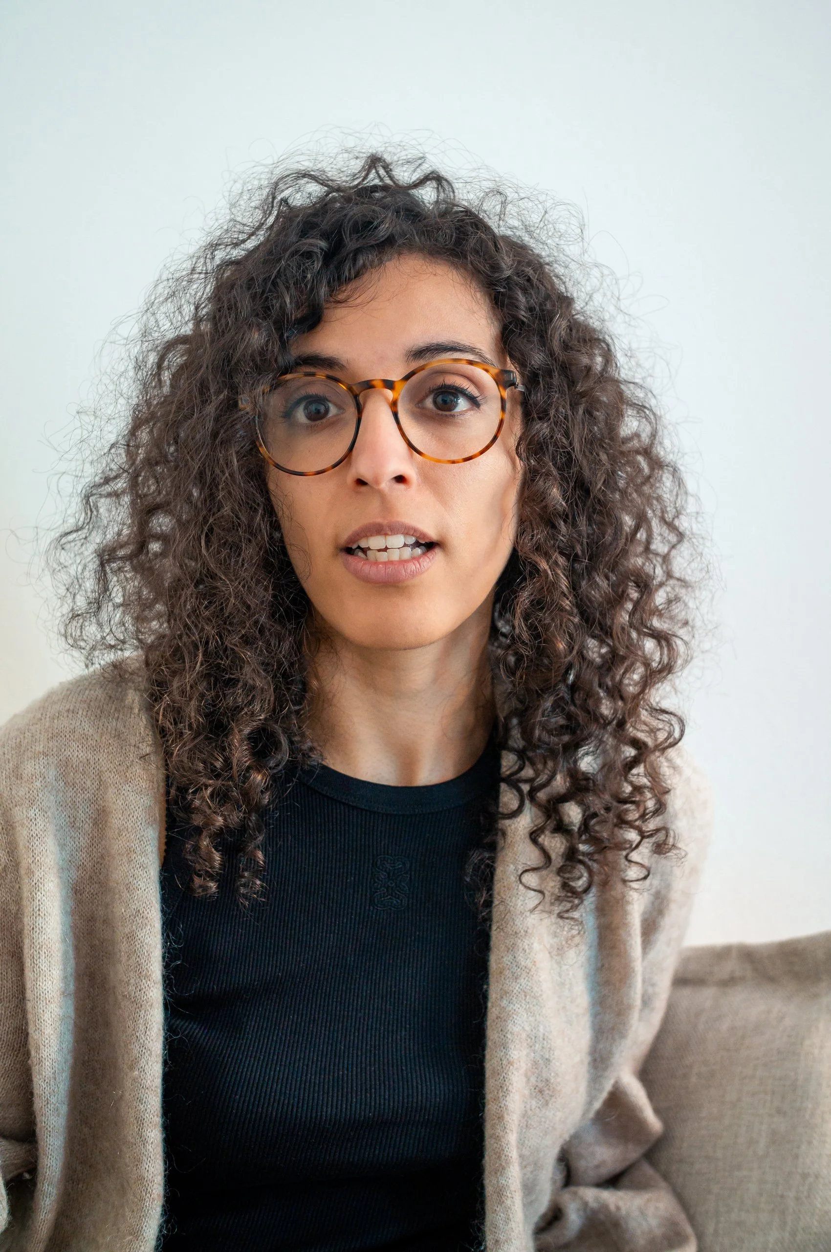 A woman with curly brown hair and glasses looking surprised or confused, wearing a black top and beige cardigan, sitting on a sofa against a white wall.
