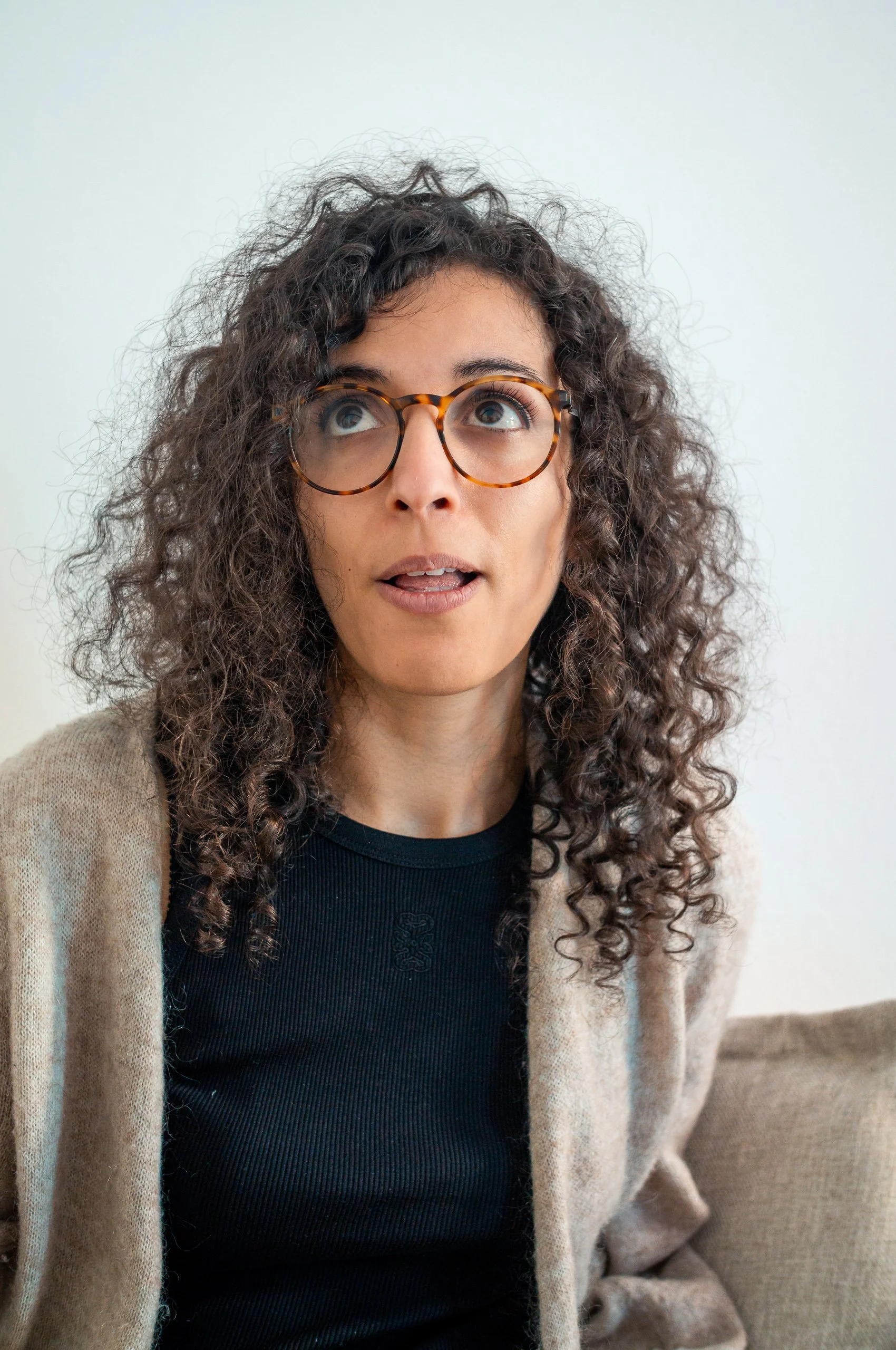 Woman with curly hair, glasses, black top, and beige cardigan sitting on a beige couch against a plain white wall.