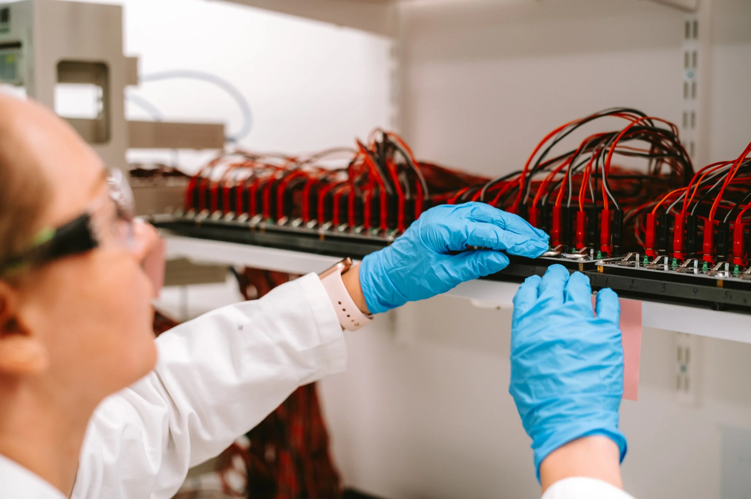 A scientist wearing blue gloves and safety glasses is working on an electronic circuit with multiple wires on a rack in a laboratory.
