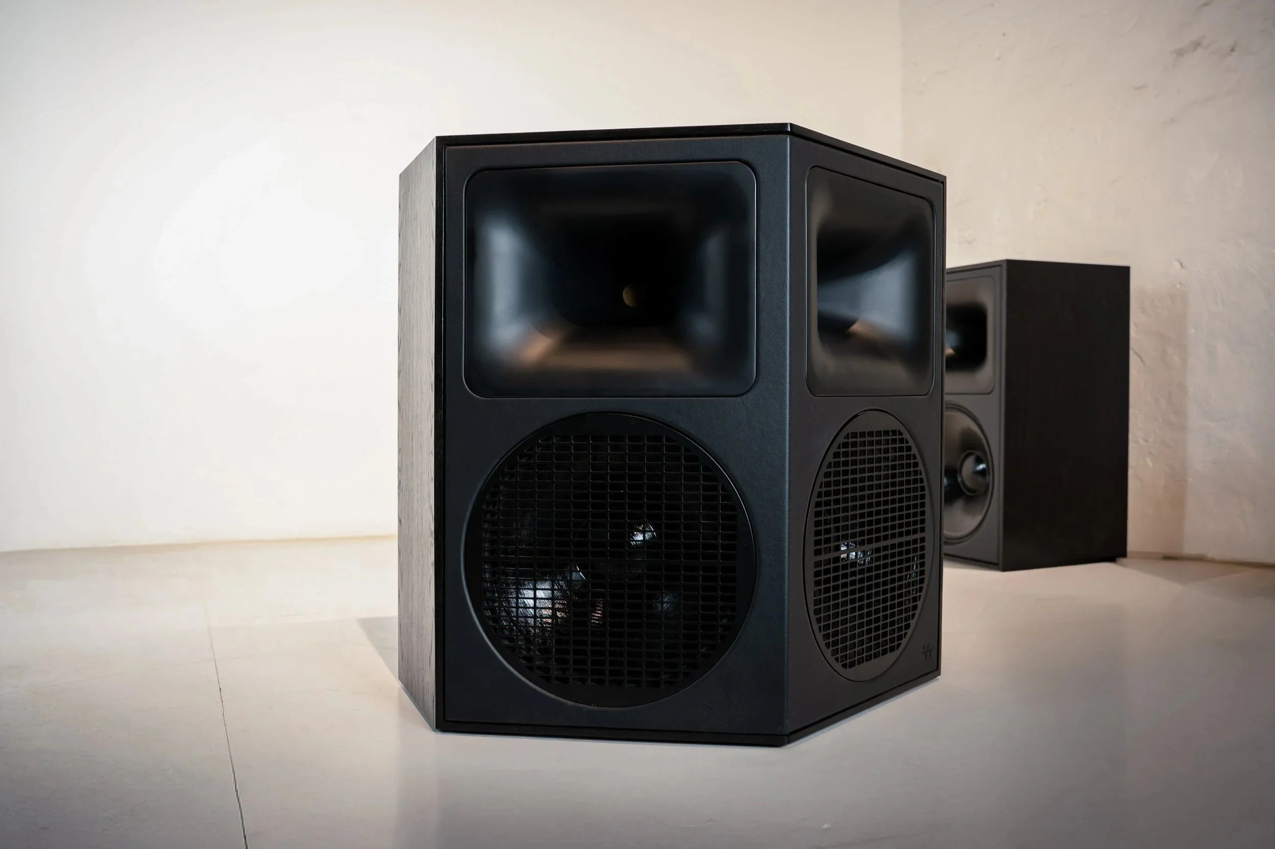 Three black speakers on a light-colored surface against a white wall, with the front two speakers prominently visible and the third partially in the background.