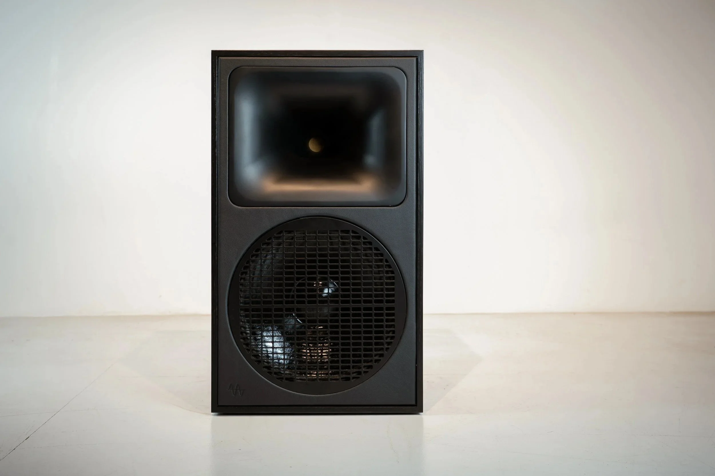 Black vintage speaker with a large circular grill and a smaller square horn on the top, standing on a white surface against a plain white wall.