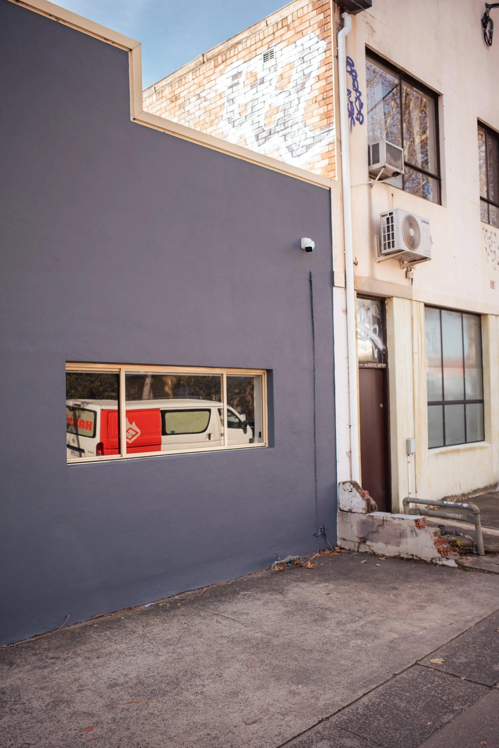 View of an urban building with a dark gray exterior wall, a small horizontal window reflecting a white van with red markings, and adjacent to a beige wall with multiple windows, air conditioning units, and graffiti. The sidewalk is concrete with a da