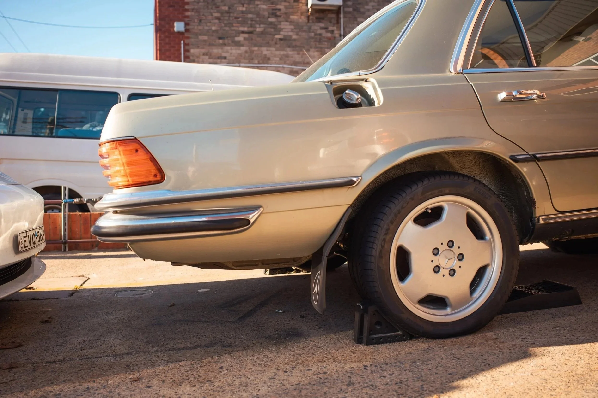A beige vintage Mercedes-Benz car parked on the street, with its front left wheel removed and placed on a jack stand.