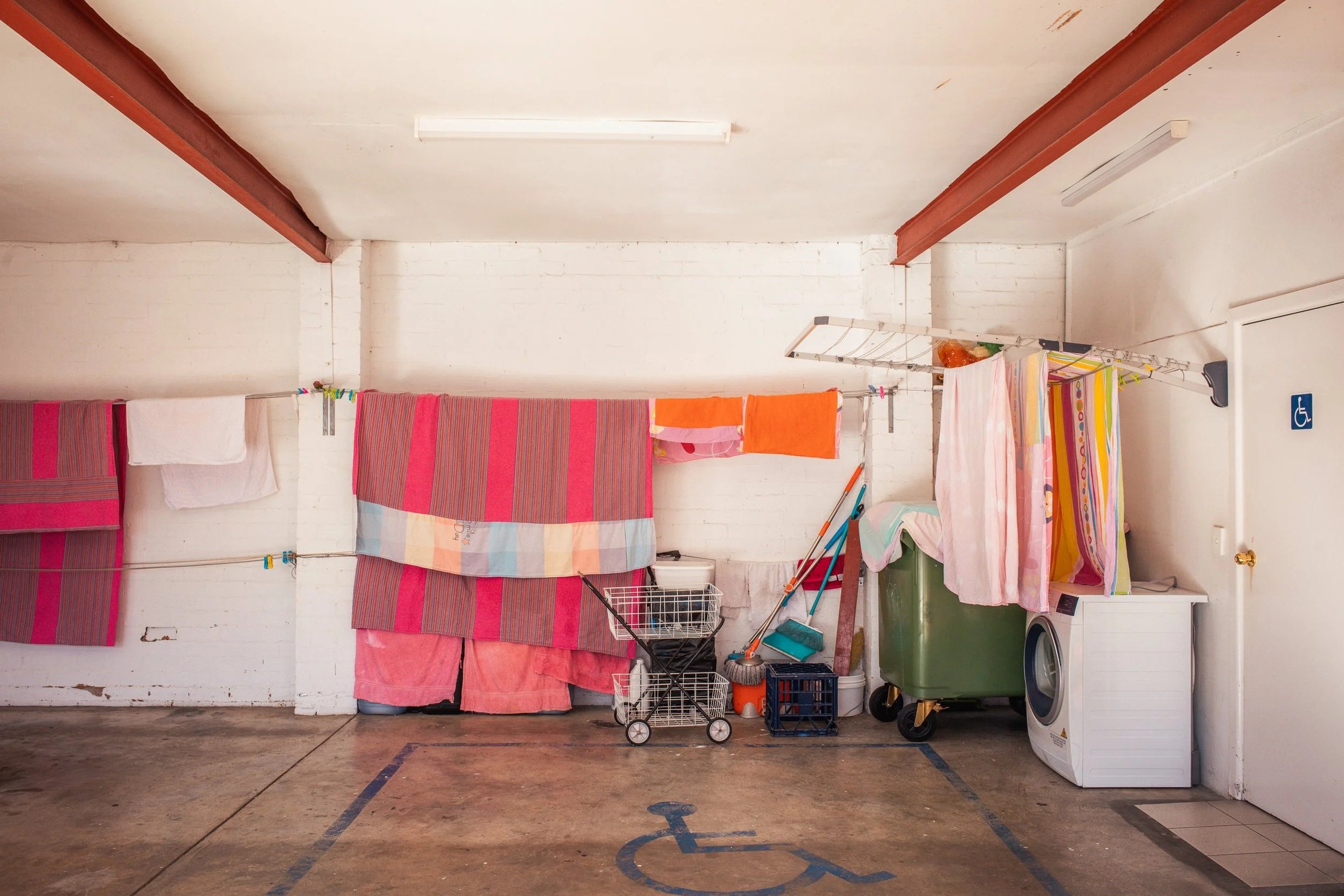 Laundry area with hanging clothes, towels, laundry cart, cleaning tools, and a washing machine in a parking space marked for disabled persons.